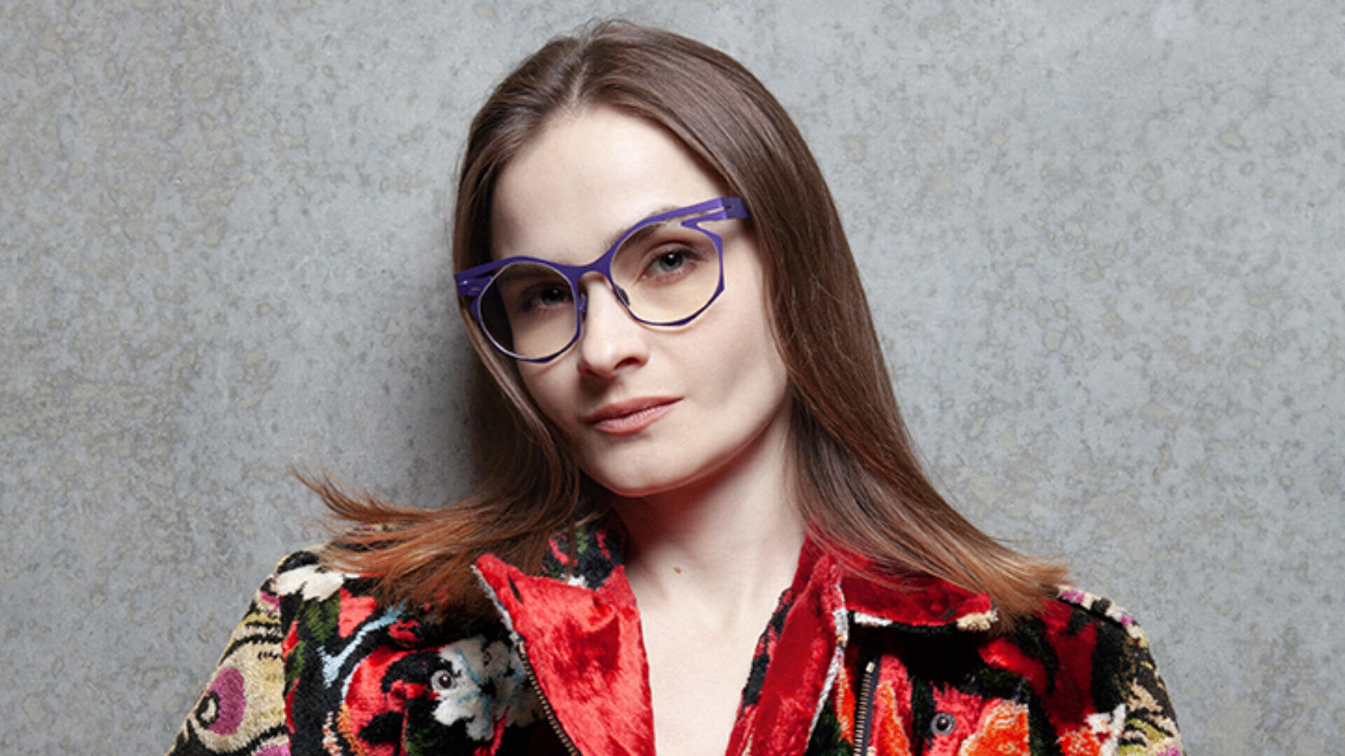 Eine Person mit glattem braunem Haar, einer lila Brille und einer bunt gemusterten Jacke steht vor einer grau strukturierten Wand und blickt selbstbewusst in die Kamera.