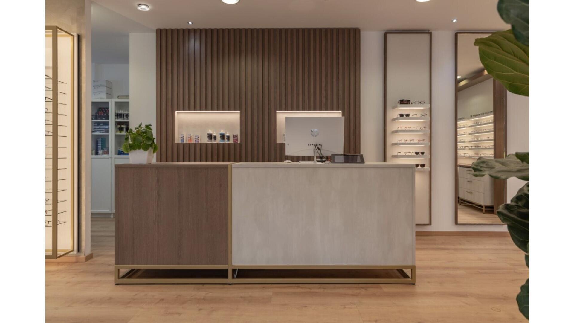 Modern optician’s reception area with a wooden and white front desk, a computer monitor, decorative wall panels, shelves displaying glasses, and a green potted plant on the left. Light wood flooring and soft lighting.