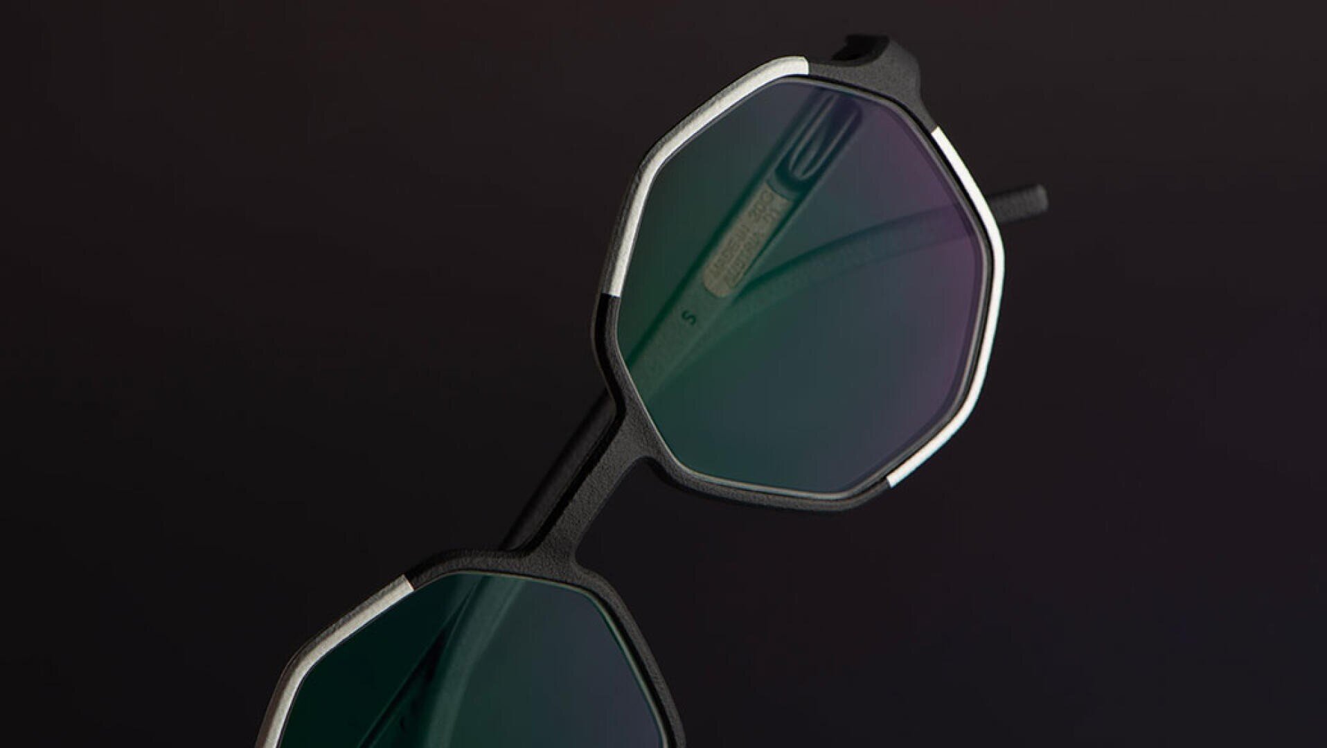 Eine Nahaufnahme einer geometrischen, achteckigen Sonnenbrille mit schwarzem Rahmen und grün getönten Gläsern vor einem dunklen Hintergrund.