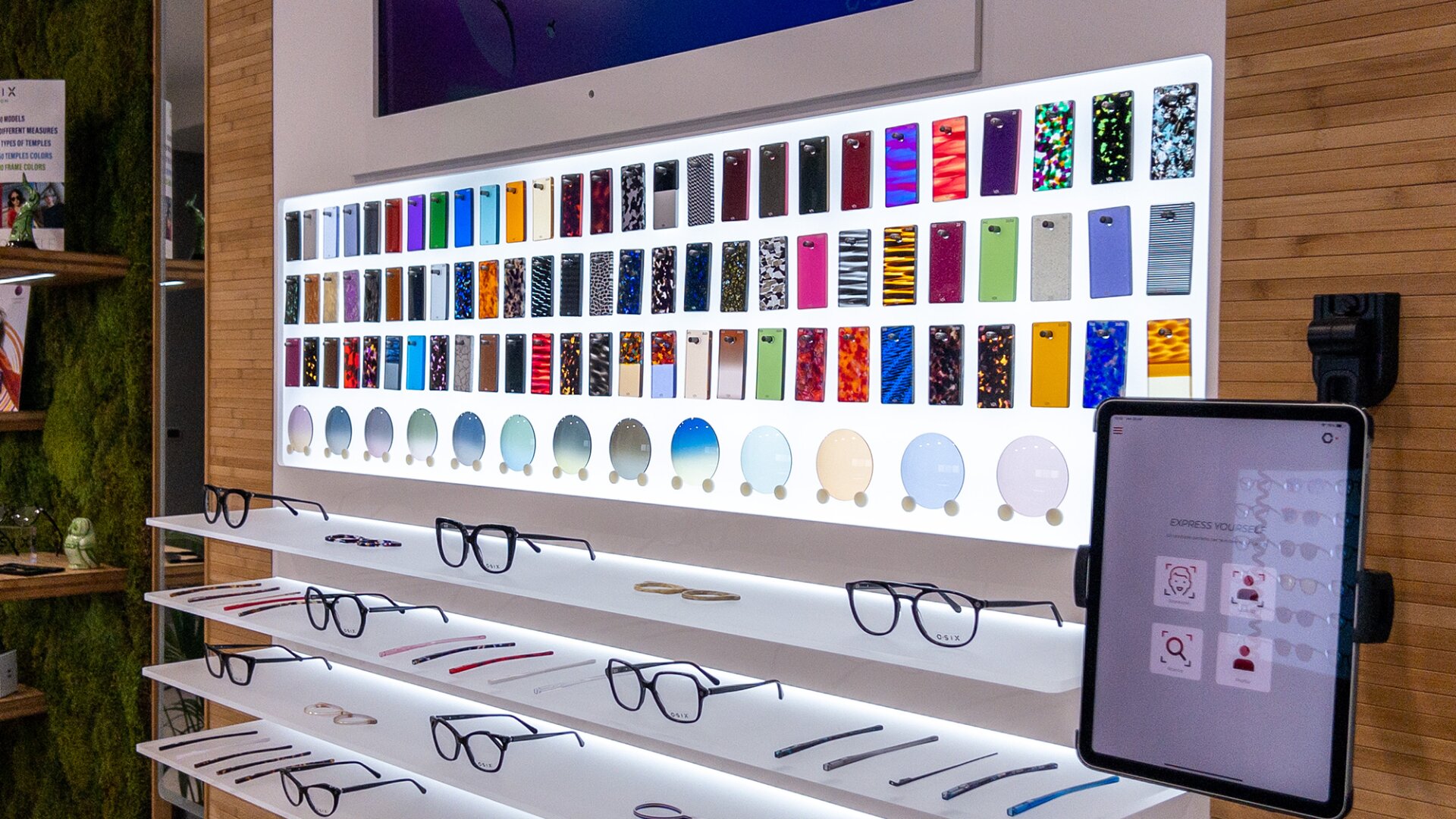 A display wall in an eyewear store shows various colorful eyeglass frame patterns and lens options above shelves with eyeglass frames. An electronic tablet is mounted on the right side.