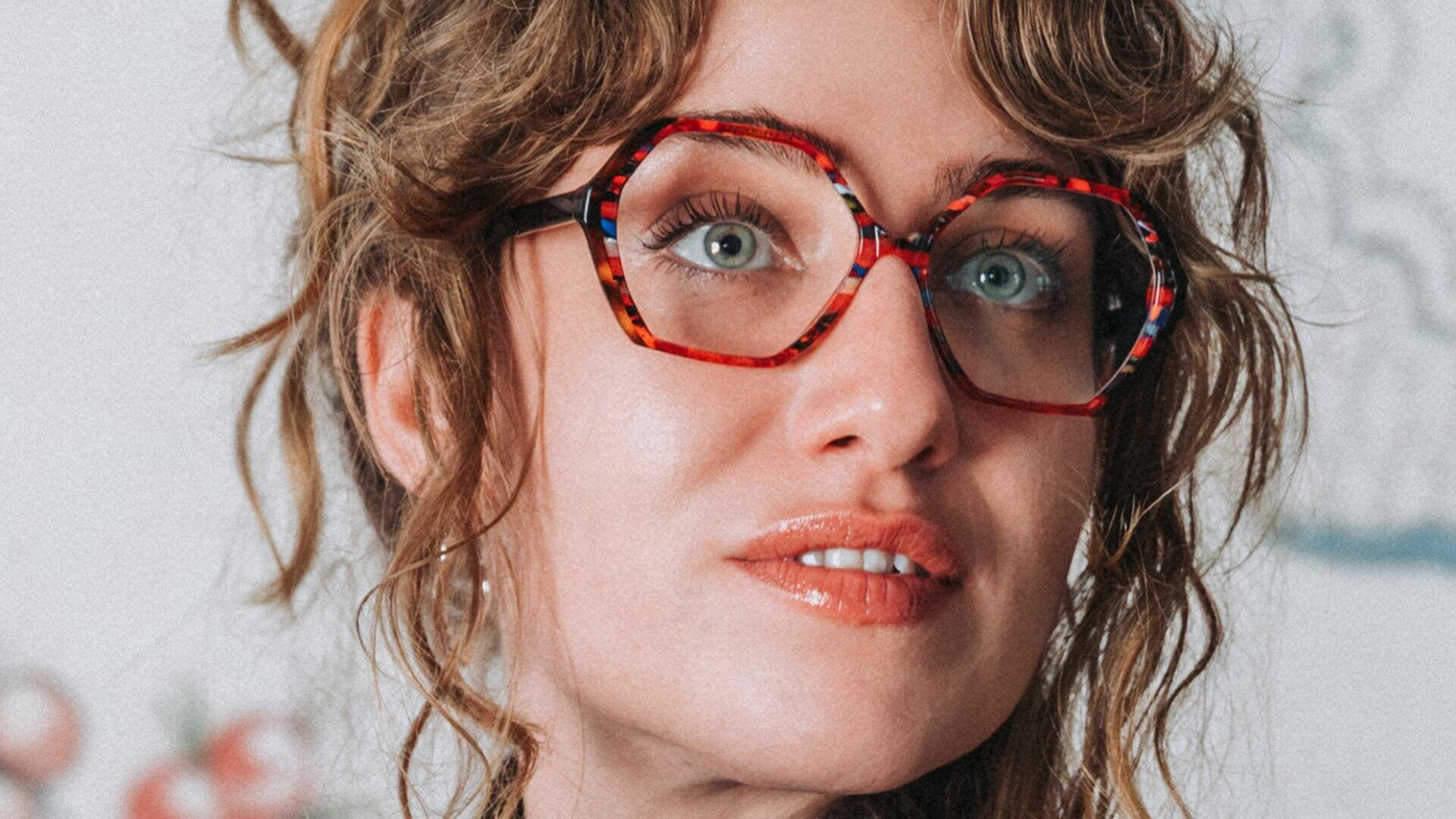 A woman with wavy light brown hair and large, colorful, round glasses looks slightly upwards and smiles gently. The background is blurred.