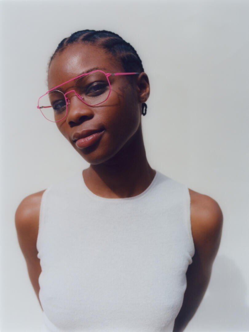 A woman with braided hair wears pink-framed glasses and a sleeveless white top, standing against a light background. She is looking at the camera with a neutral expression and her arms are behind her back.