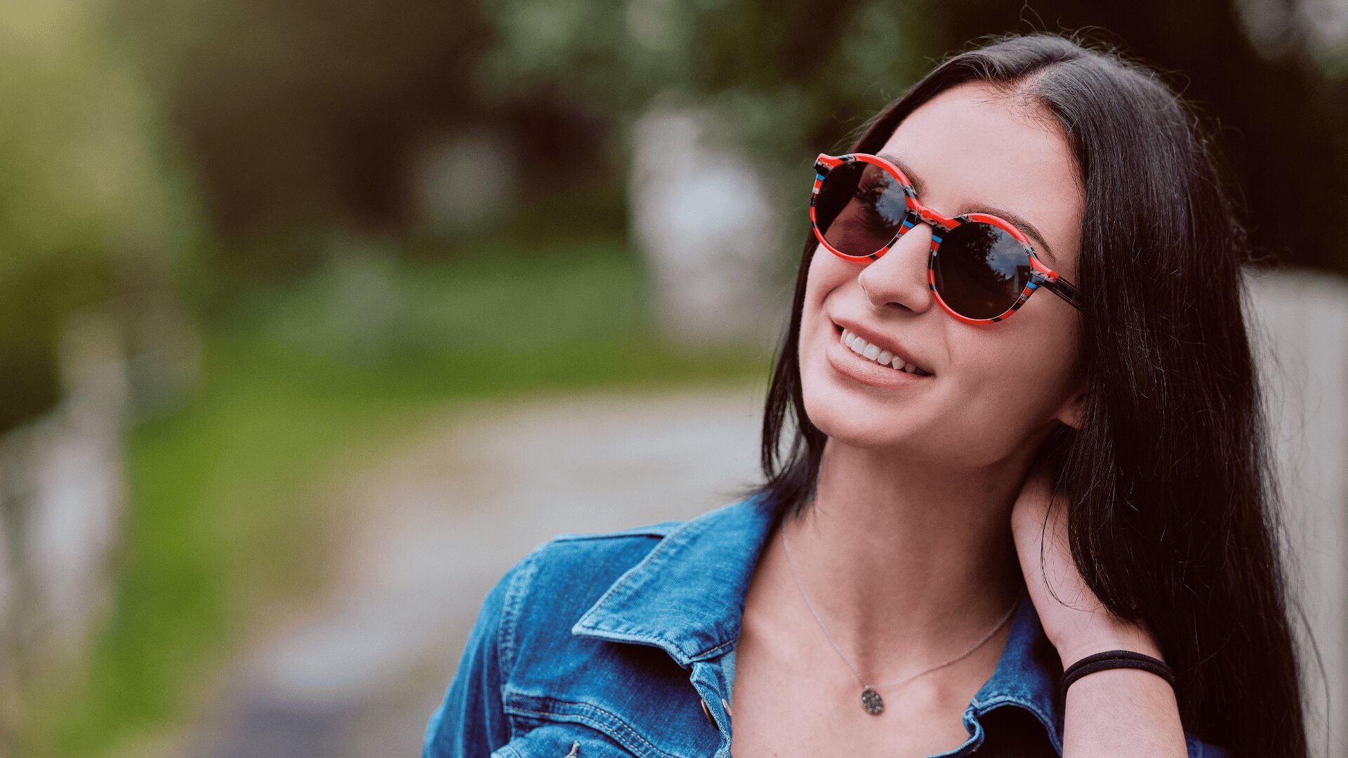 Eine junge Frau mit langen dunklen Haaren, die eine rote Sonnenbrille und eine Jeansjacke trägt, lächelt, während sie ihren Kopf neigt und ihren Nacken berührt, vor einem verschwommenen grünen Hintergrund im Freien.