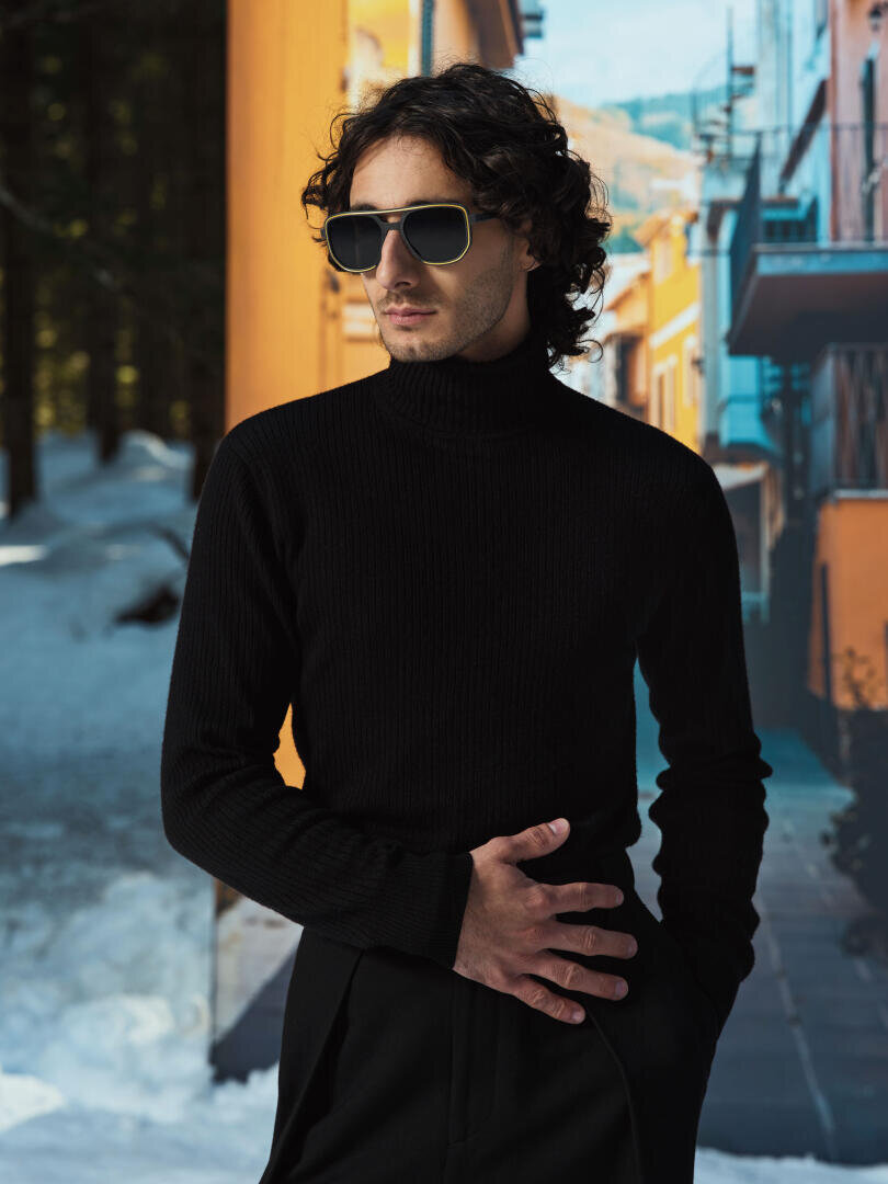 Ein Mann mit dunkler Sonnenbrille und schwarzem Rollkragenpullover steht im Freien, eine Hand in der Taille. Im Hintergrund sind Schnee und bunte Gebäude zu sehen.
