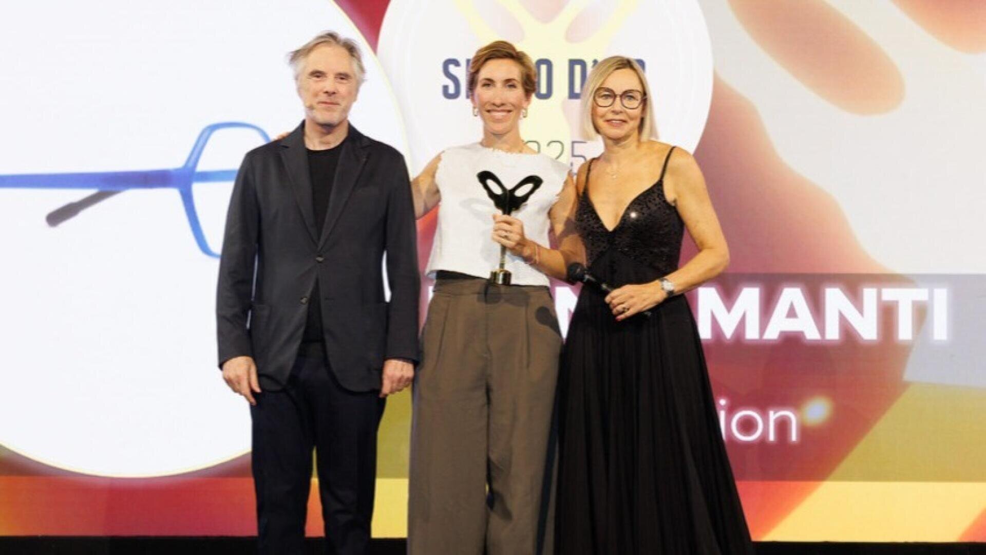 Drei Personen stehen bei einer Preisverleihung auf der Bühne. Die Frau in der Mitte hält eine Trophäe. Im Hintergrund ist ein bunter Bildschirm mit Text und Grafiken zu sehen. Alle drei Personen lächeln und sind förmlich gekleidet.