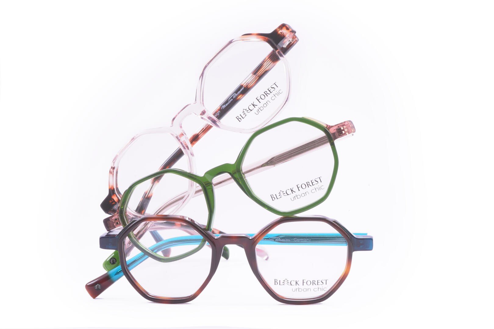 Drei Brillenpaare der Marke Black Forest Urban Chic – eine klare, eine grüne und eine in Schildpattoptik – sind diagonal vor einem weißen Hintergrund gestapelt.