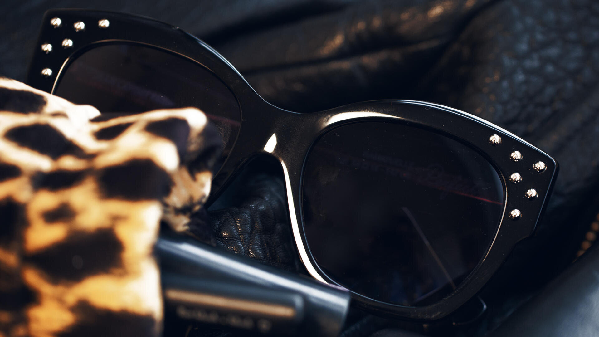 Nahaufnahme einer schwarzen Cat-Eye-Sonnenbrille mit kleinen silbernen Nieten am Rahmen, die auf schwarzem Ledermaterial neben einem Stoff mit Leopardenmuster und einem dunklen zylindrischen Objekt ruht.