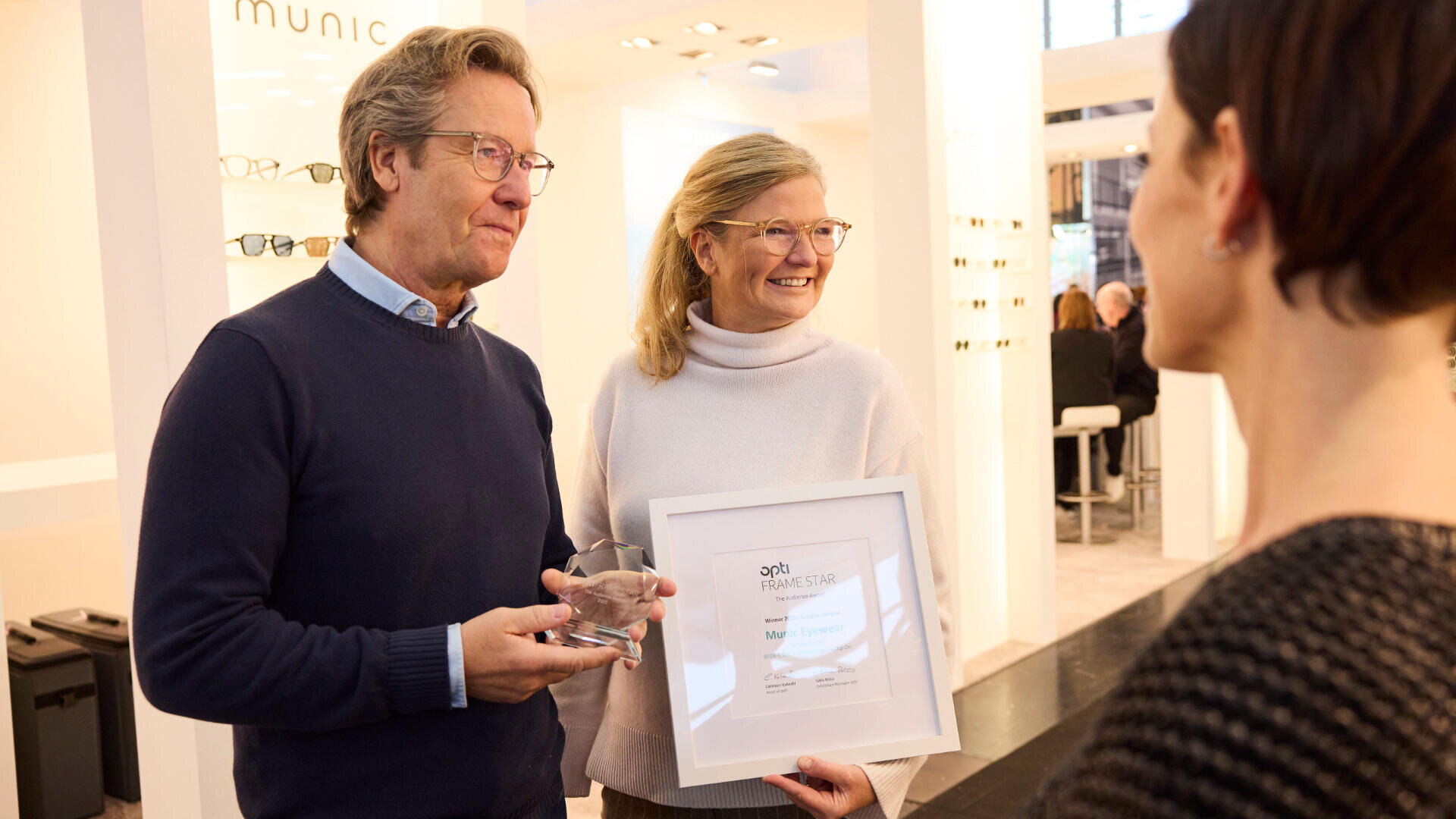 Zwei lächelnde Personen halten eine Auszeichnung und ein gerahmtes Zertifikat in den Händen, während sie sich in einem gut beleuchteten Innenraum, möglicherweise bei einer Veranstaltung oder Ausstellung, mit einer dritten Person unterhalten. Im Hintergrund sind Gläser ausgestellt.