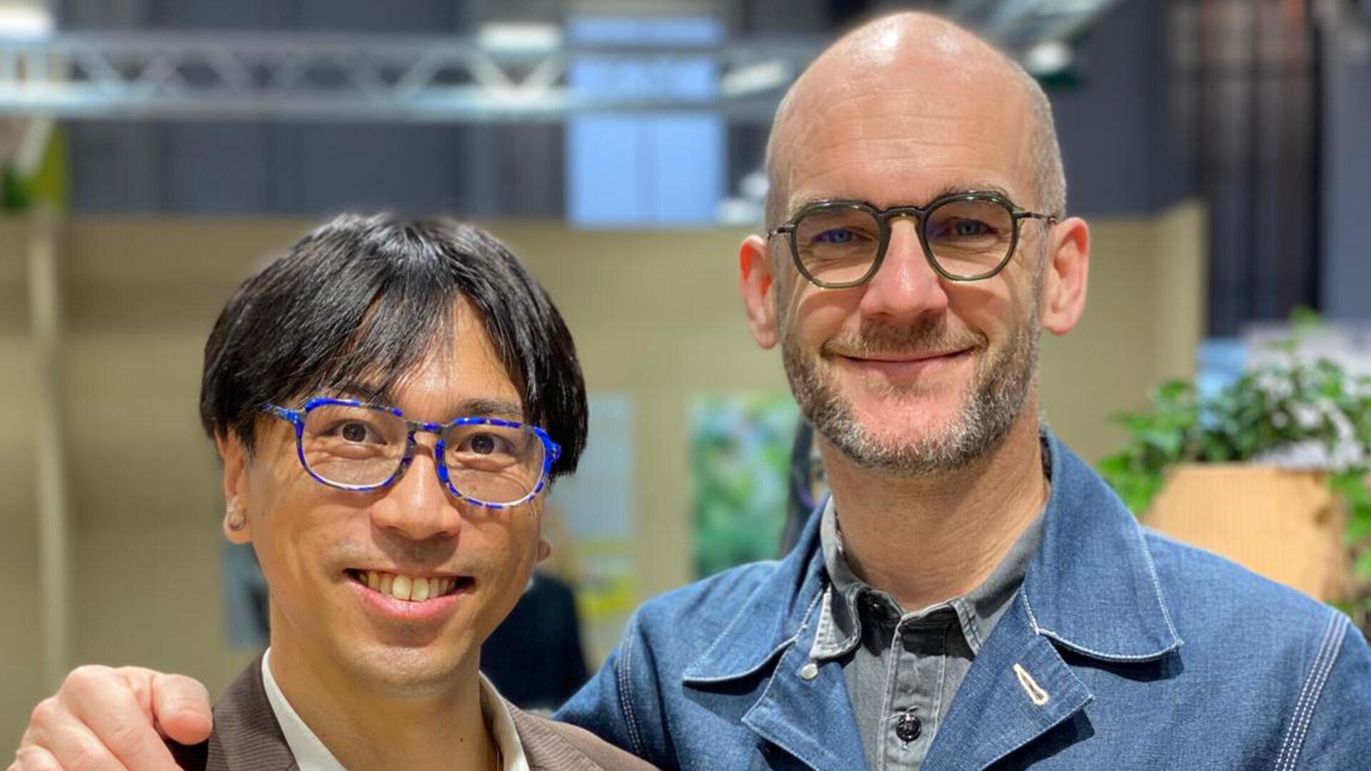 Zwei lächelnde Männer mit Brille posieren zusammen in einem Innenbereich. Der eine hat kurze dunkle Haare und eine blaue Brille, der andere ist kahlköpfig, hat einen Bart und eine runde Brille. Beide tragen Jacken. Der Hintergrund ist leicht unscharf.