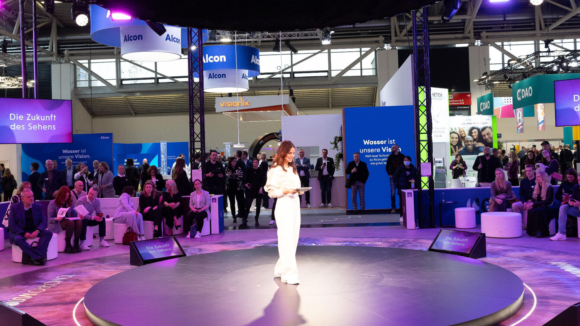 Eine Frau in Weiß steht auf einer runden Bühne und spricht zu einem Publikum bei einer Technologieausstellung. Hinter ihr befinden sich blaue und violette Displays und um die Bühne herum sitzende und stehende Menschen.