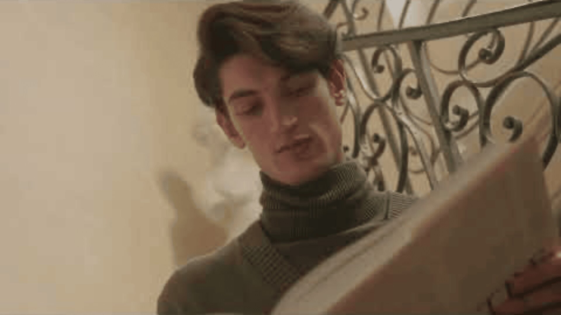 Ein junger Mann mit welligem dunklem Haar und einem Rollkragenpullover steht im Innenbereich und blickt auf ein großes, aufgeschlagenes Buch. Im Hintergrund ist ein dekoratives Geländer zu sehen und es herrscht eine warme Beleuchtung.