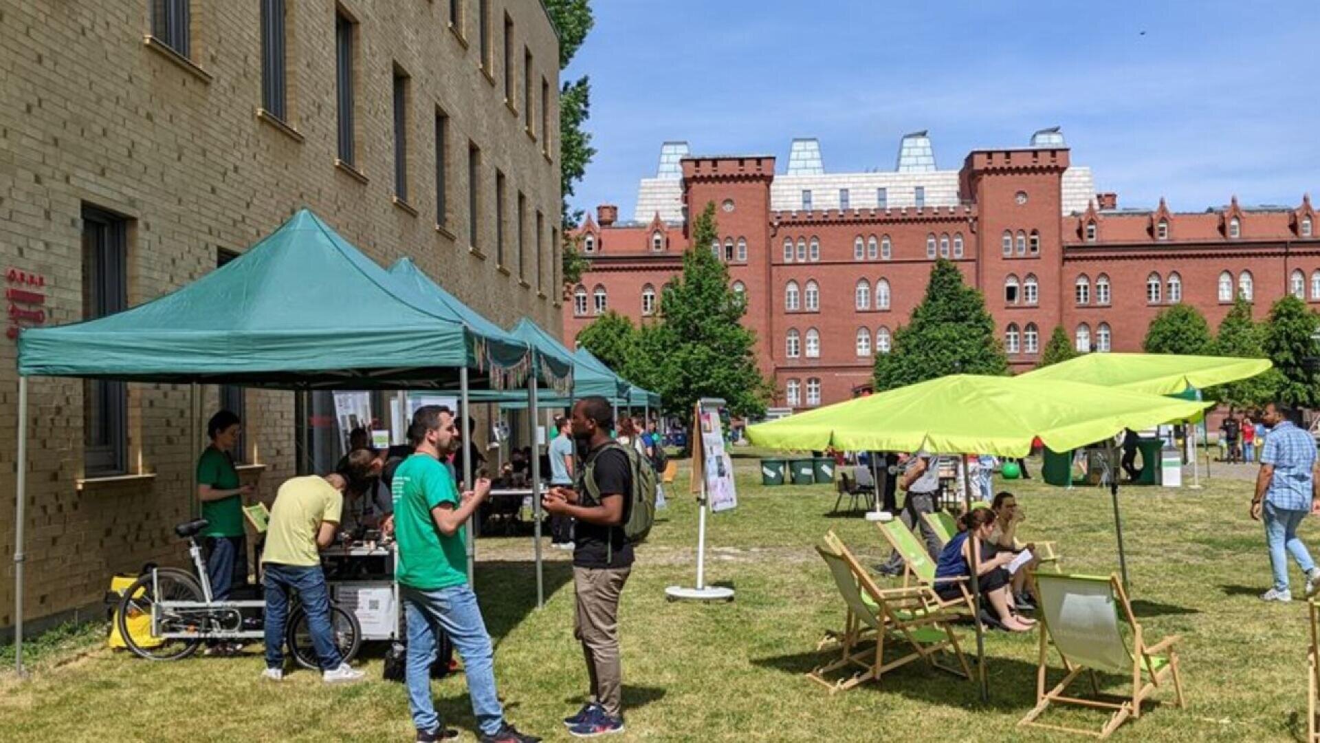 People gather outdoors near a large brick building with green tents and umbrellas set up. Some are talking, others sit on chairs in the sun. The scene appears to be a community or campus event on a grassy lawn.