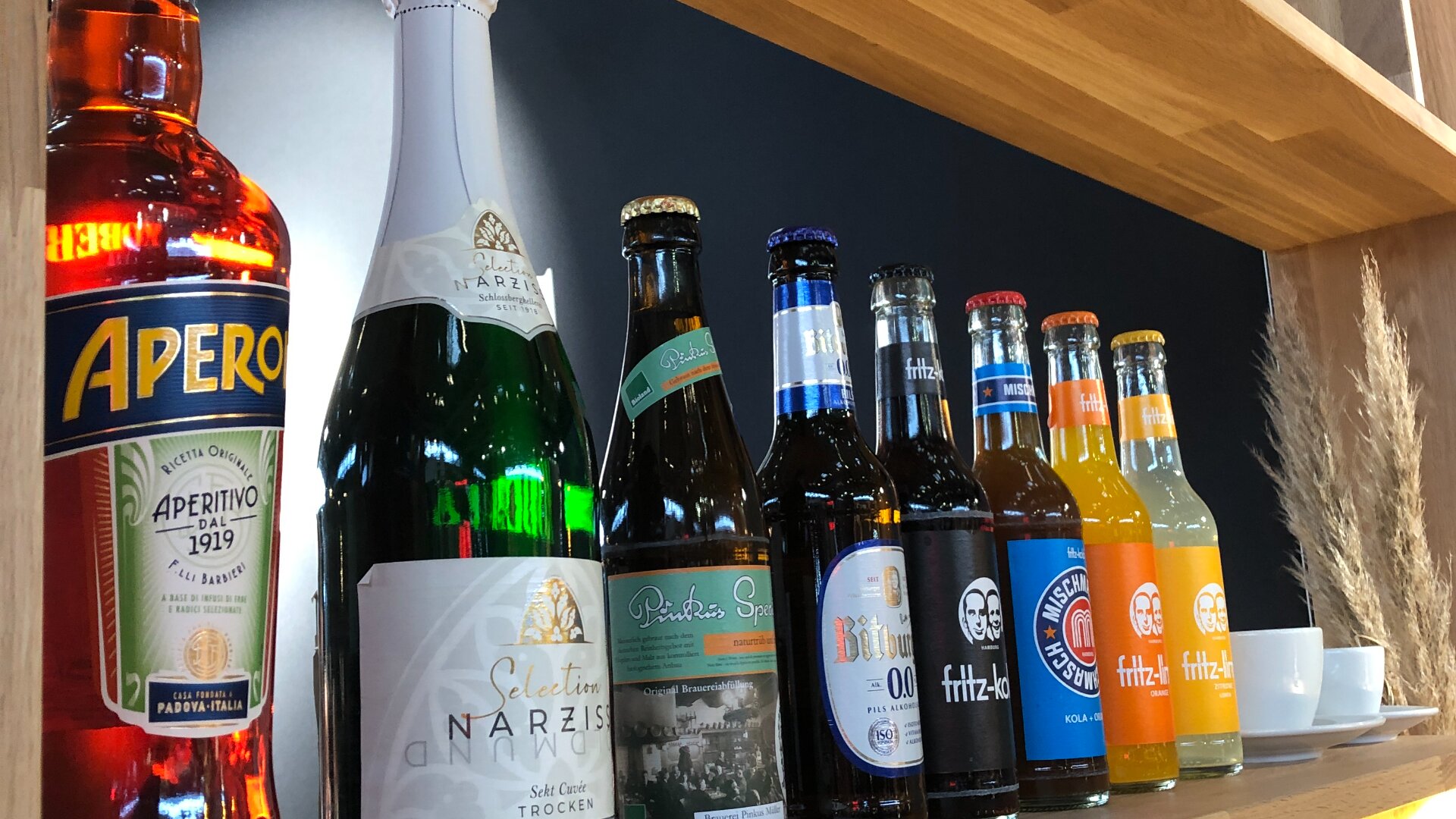 Auf einem Holzregal stehen Flaschen mit Aperol, Sekt, verschiedenen Bieren und bunten Softdrinks vor dunklem Hintergrund. Die Getränke sind ordentlich aufgereiht und zeigen ihre Etiketten.