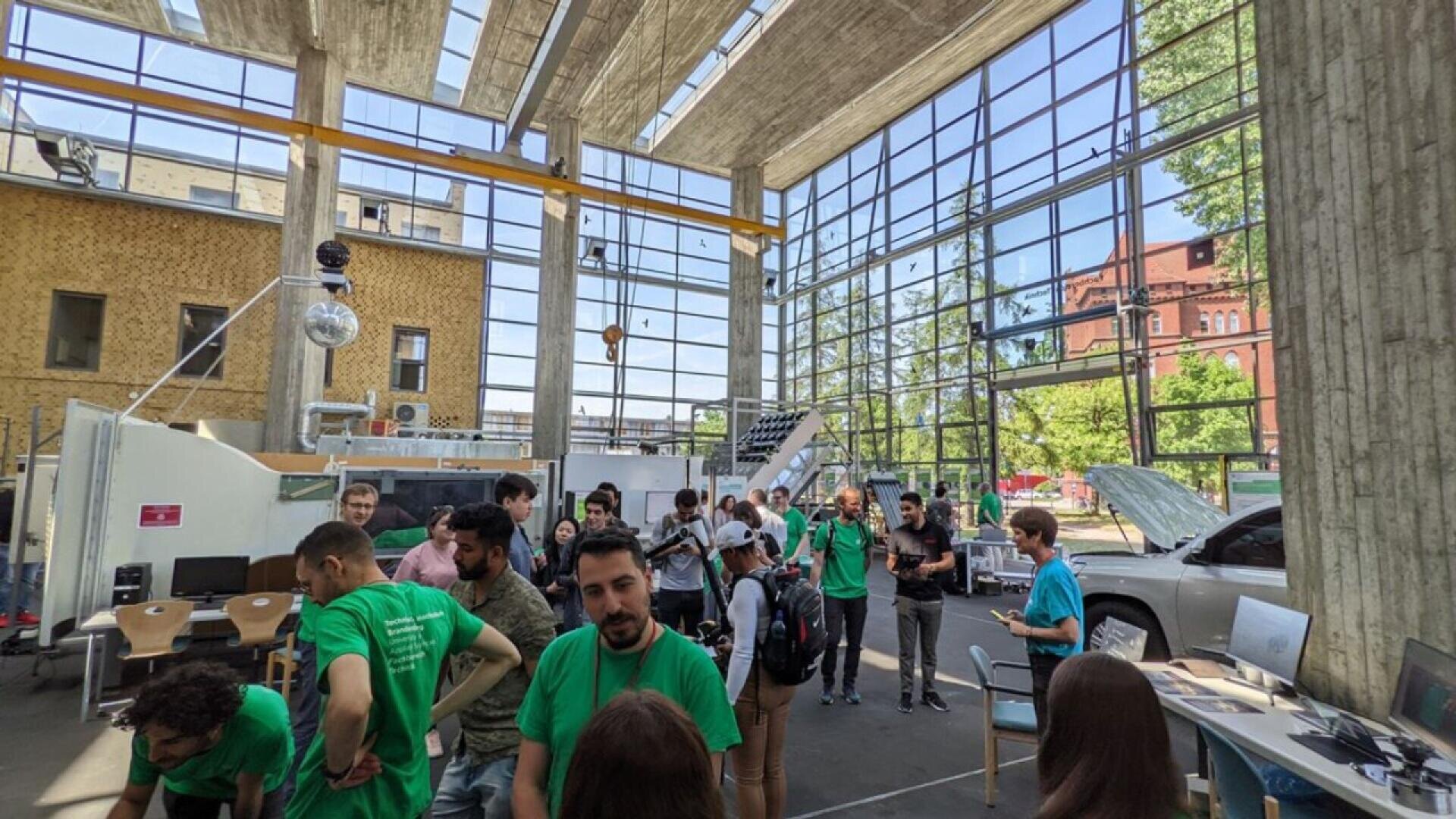A group of people, some in green shirts, gather and interact in a spacious, sunlit room with large glass windows, concrete beams, and technology exhibits visible in the background.