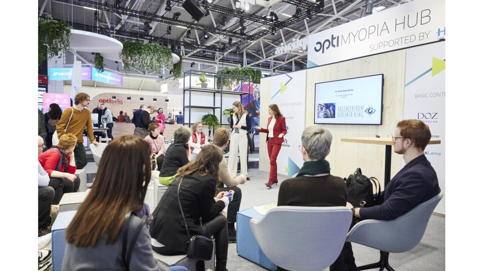 A group of people sits and listens to a presenter speaking at the Myopia Hub booth during an indoor event or conference, with a screen displaying a slide and banners in the background.