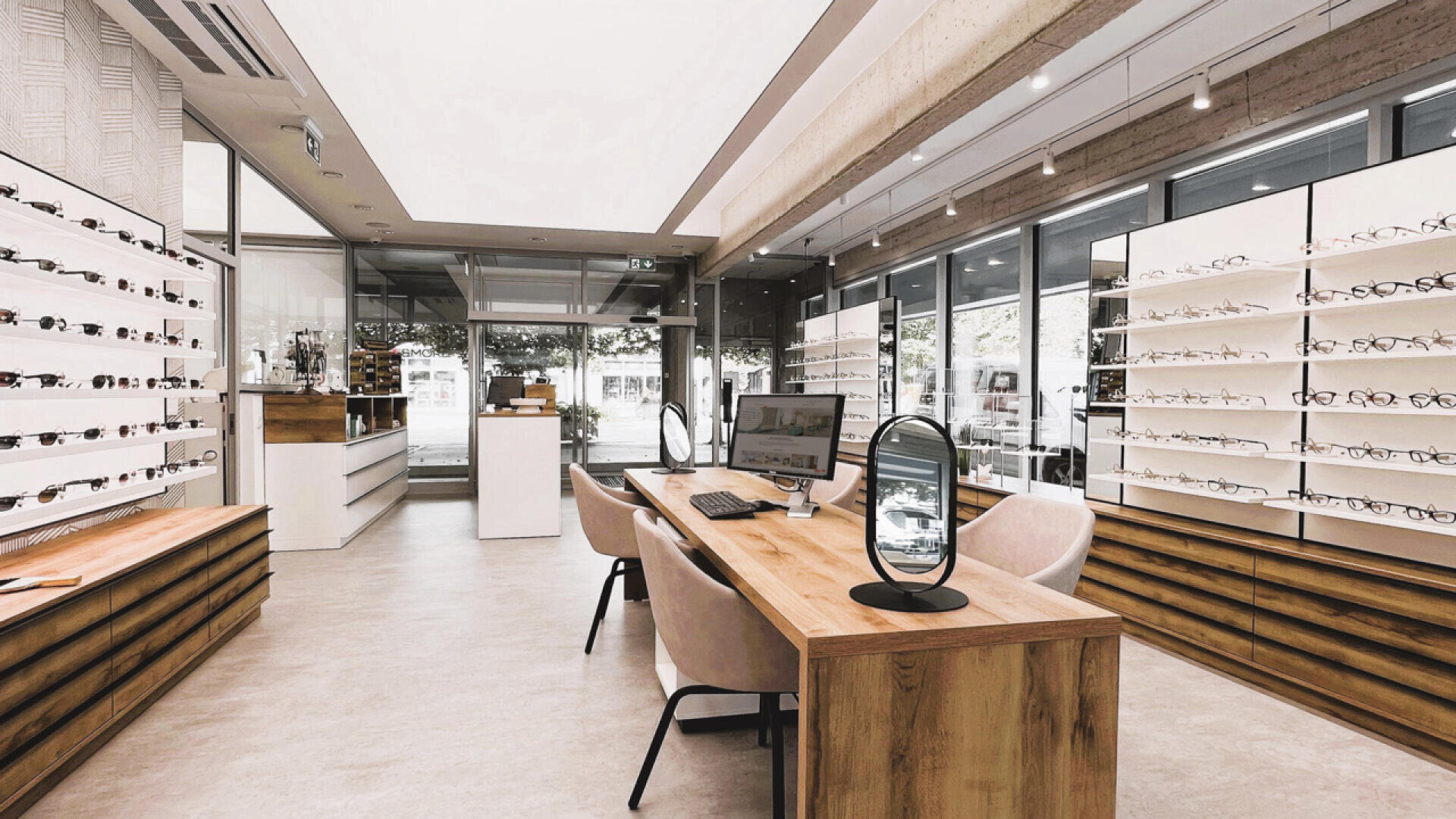 A modern optician’s store with wooden desks, display shelves filled with eyeglasses, two chairs at each desk, computers, and large windows letting in natural light.