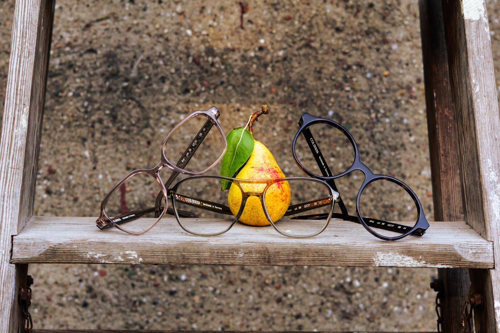 Eine gelbe Birne mit einem grünen Blatt liegt auf einem verwitterten Holzregal, flankiert von drei Brillen mit verschiedenfarbigen Rahmen. Der Hintergrund ist eine raue, gesprenkelte graue Fläche.