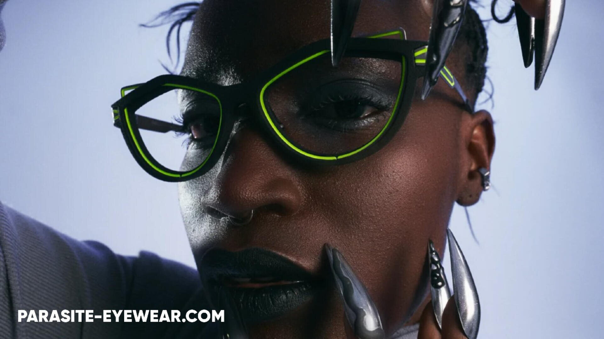 Eine Person trägt eine schwarz-neongrüne Brille, dramatisches Make-up und dunklen Lippenstift. Ihre Fingernägel sind metallisch und krallenartig. In der unteren linken Ecke erscheint der Text PARASITE-EYEWEAR.COM.