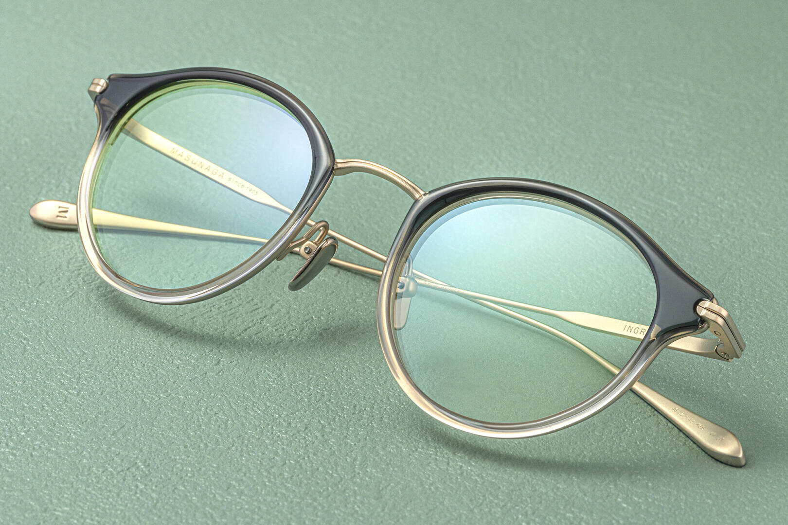 Round eyeglasses with thin gold metal arms and translucent blue-tinted lenses are resting on a textured green surface. The frames have a gradient design, blending black on top and clear on the bottom.