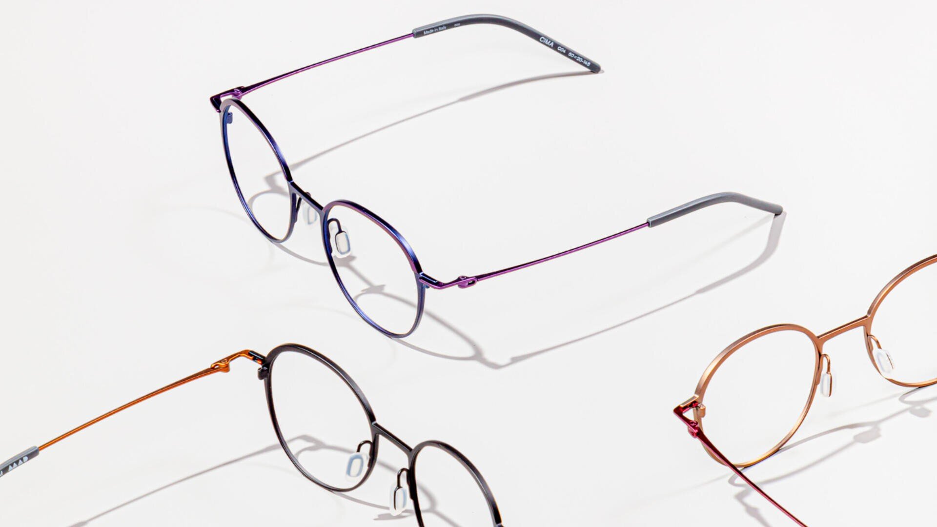 Three pairs of round eyeglasses with thin metal frames in different colors—purple, black, and copper—are arranged on a white surface, casting soft shadows.