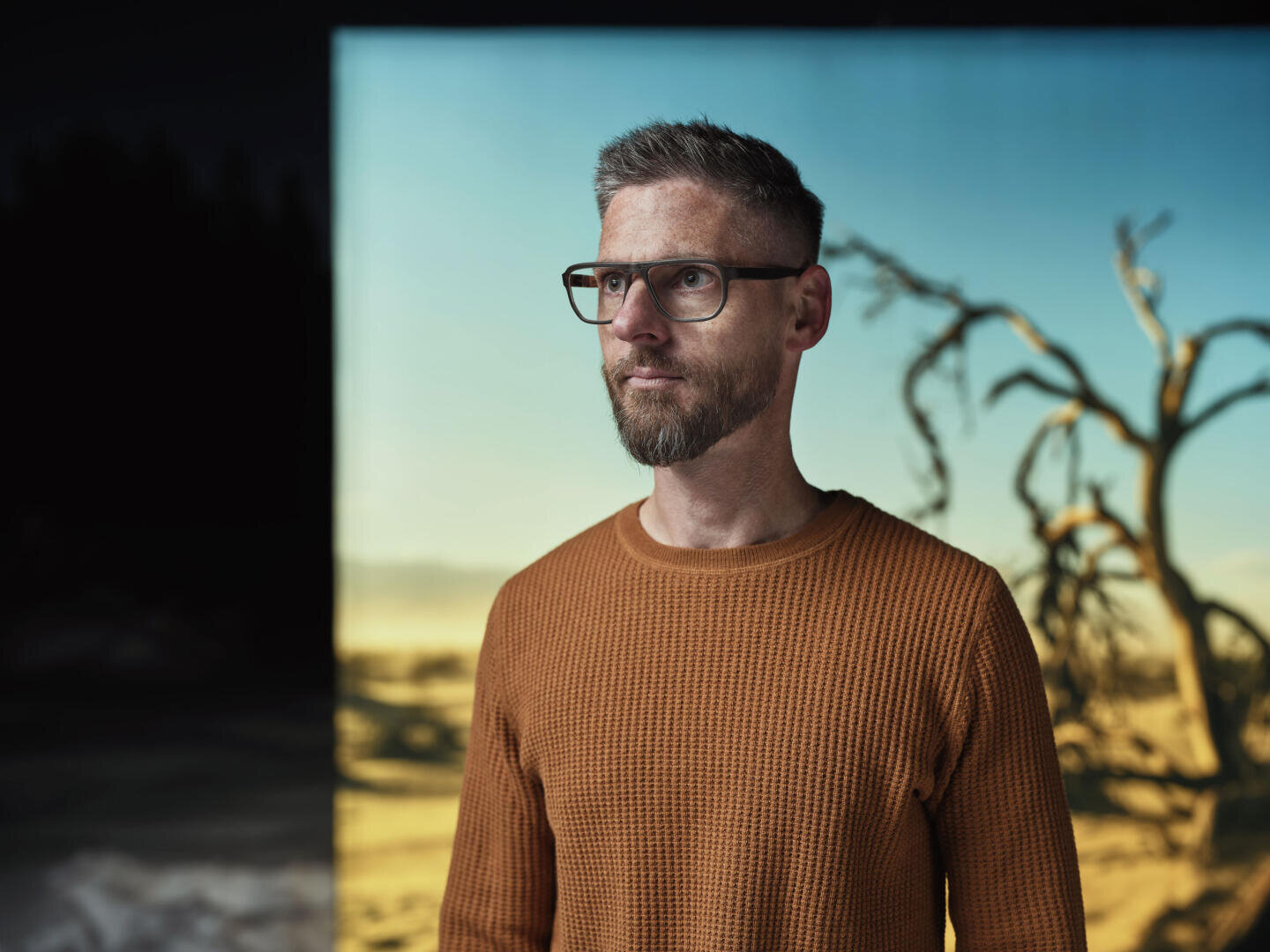 Ein Mann mit kurzen Haaren, Brille und Bart, der einen strukturierten orangefarbenen Pullover trägt, steht vor einem Hintergrund, der eine trockene Landschaft mit einem blattlosen Baum unter einem blauen Himmel zeigt.