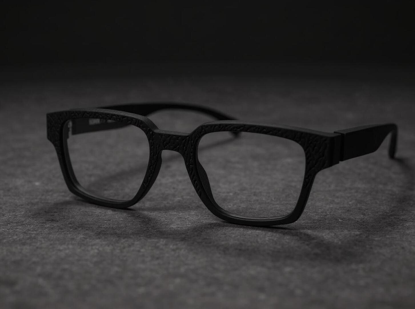 A pair of black rectangular glasses with textured frames that rest on a dark, textured surface against a black background.