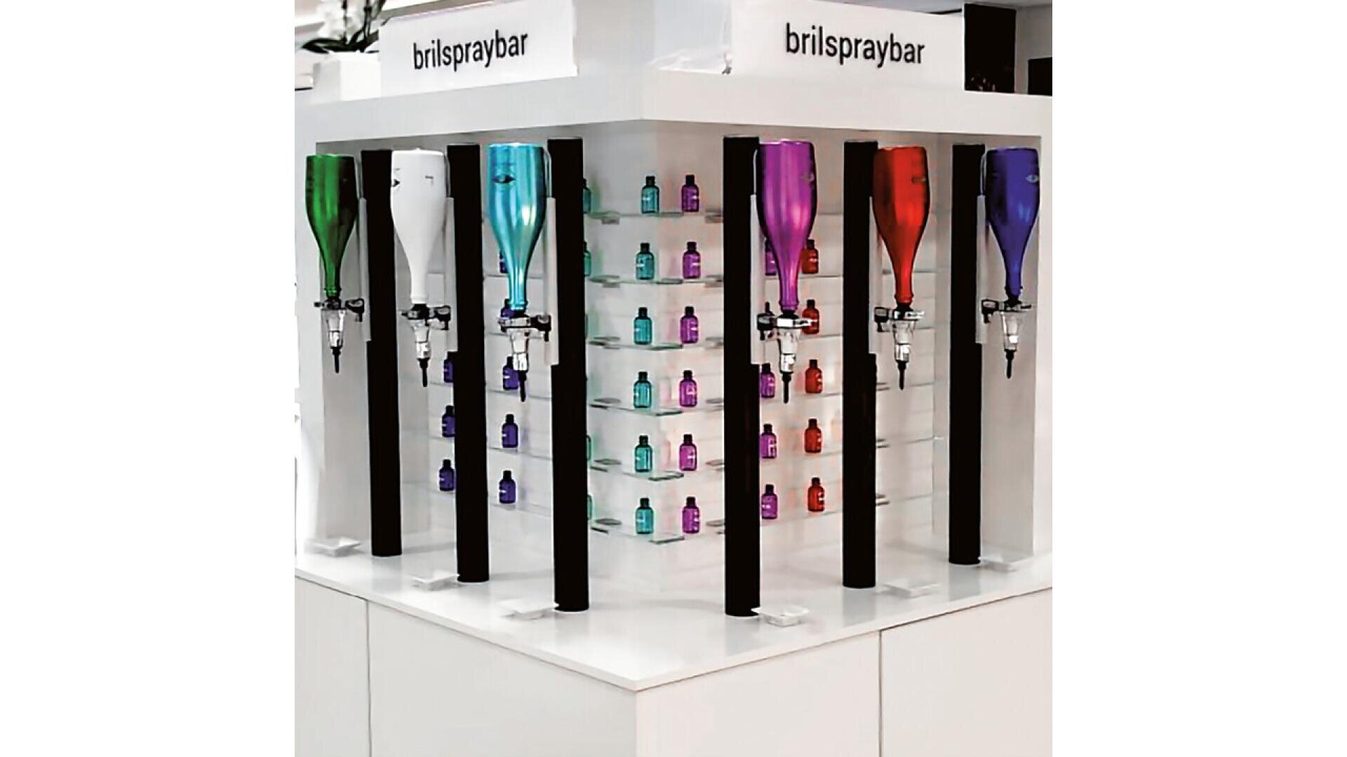 A display stand labeled brilspraybar featuring six large, colorful spray bottles and shelves with small bottles in various colors, arranged neatly in rows.