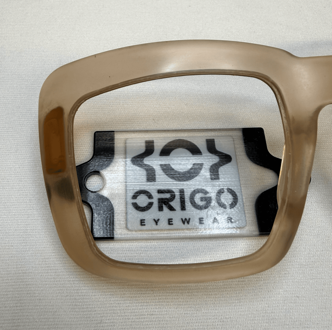 Ein beiger Brillenrahmen, der über eine Karte mit dem Logo und dem Text ORIGO EYEWEAR auf einer weißen Fläche gehalten wird. Das Brillenglas vergrößert und verdeutlicht den Text und das Logo darunter.