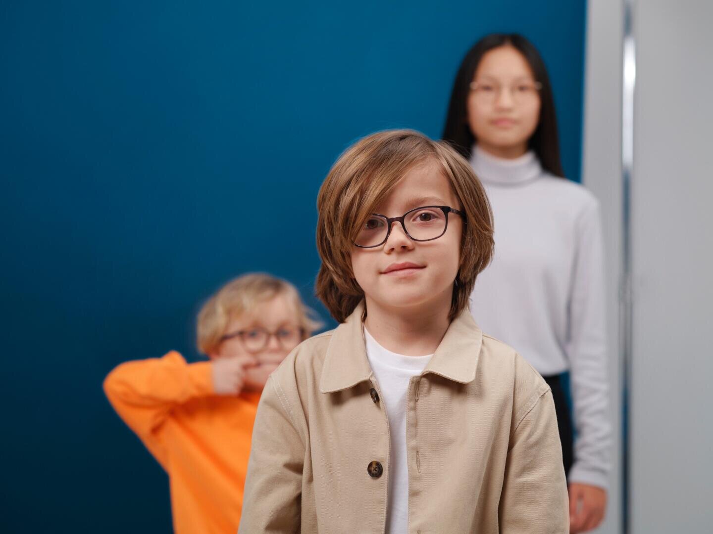 Drei Kinder stehen in einem Haus vor einem blauen Hintergrund. Ein Junge mit Brille steht vorne und blickt in die Kamera; hinter ihm macht ein Kind in Orange ein Gesicht, und ein anderes Kind in Weiß steht weiter hinten, leicht unscharf.