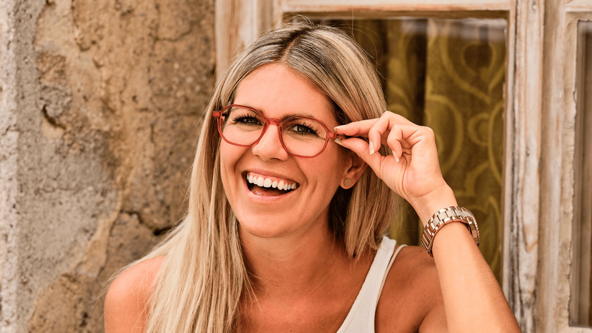 Eine lächelnde Frau mit langem blondem Haar und roter Brille sitzt vor einer verwitterten Wand und einem Fenster, trägt ein ärmelloses Oberteil und eine Uhr und hält ihre Brille mit einer Hand.