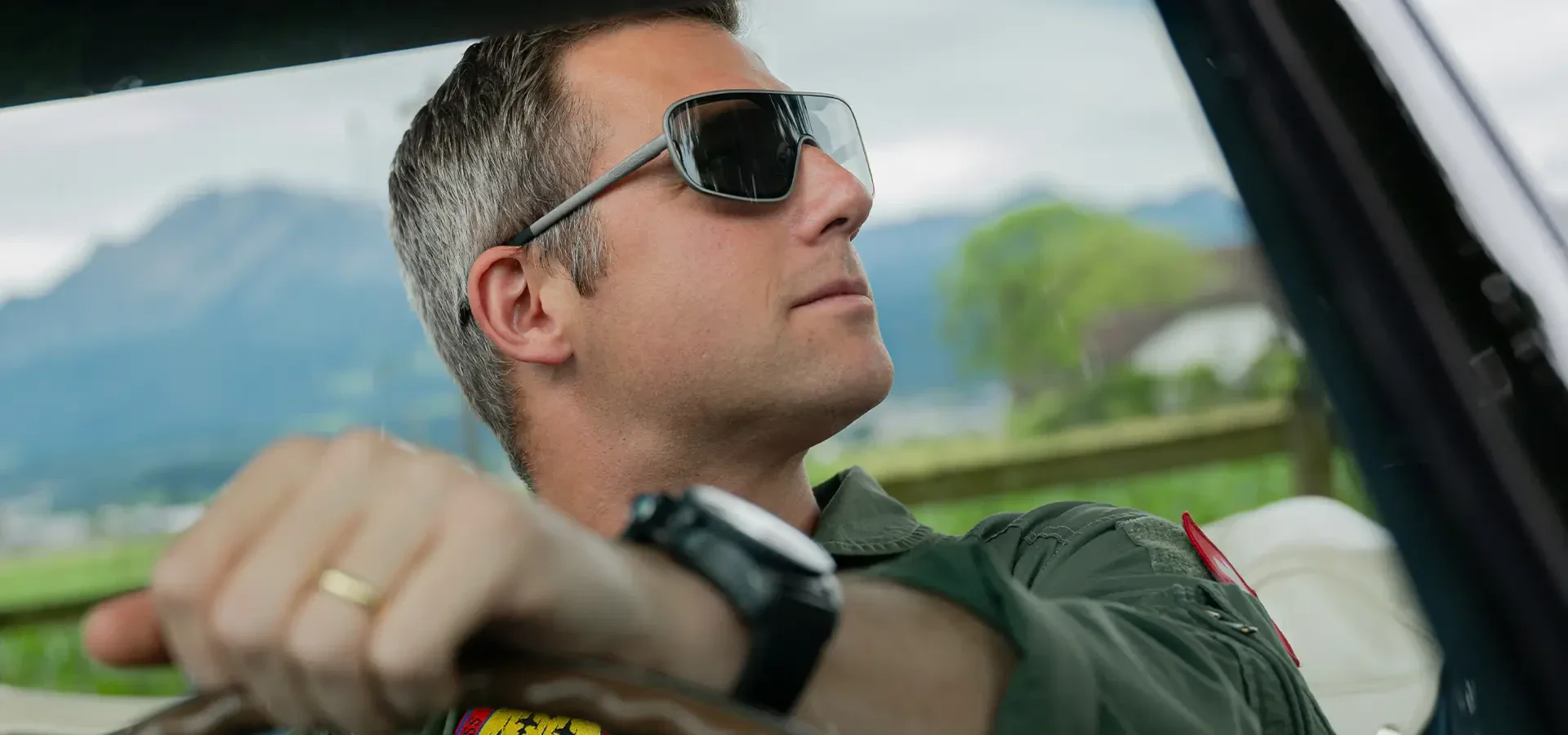 Ein Mann mit Sonnenbrille und Uhr fährt ein Auto und schaut aus dem Fenster. Er ist mit einer grünen Jacke bekleidet und hat kurze Haare. Im Hintergrund sind verschwommene grüne Flächen und ferne Berge zu sehen.