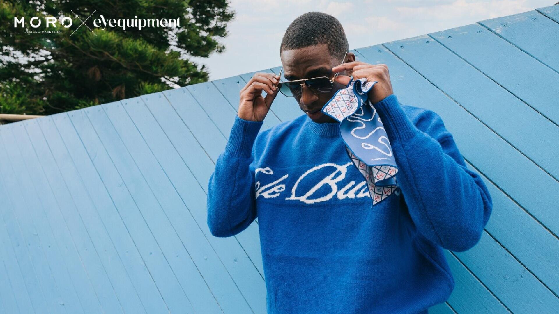 A man in a bright blue sweater adjusts his sunglasses while holding a patterned cloth. He stands in front of a blue wooden wall outdoors, with logos MORD and eleequiipment in the top left corner.