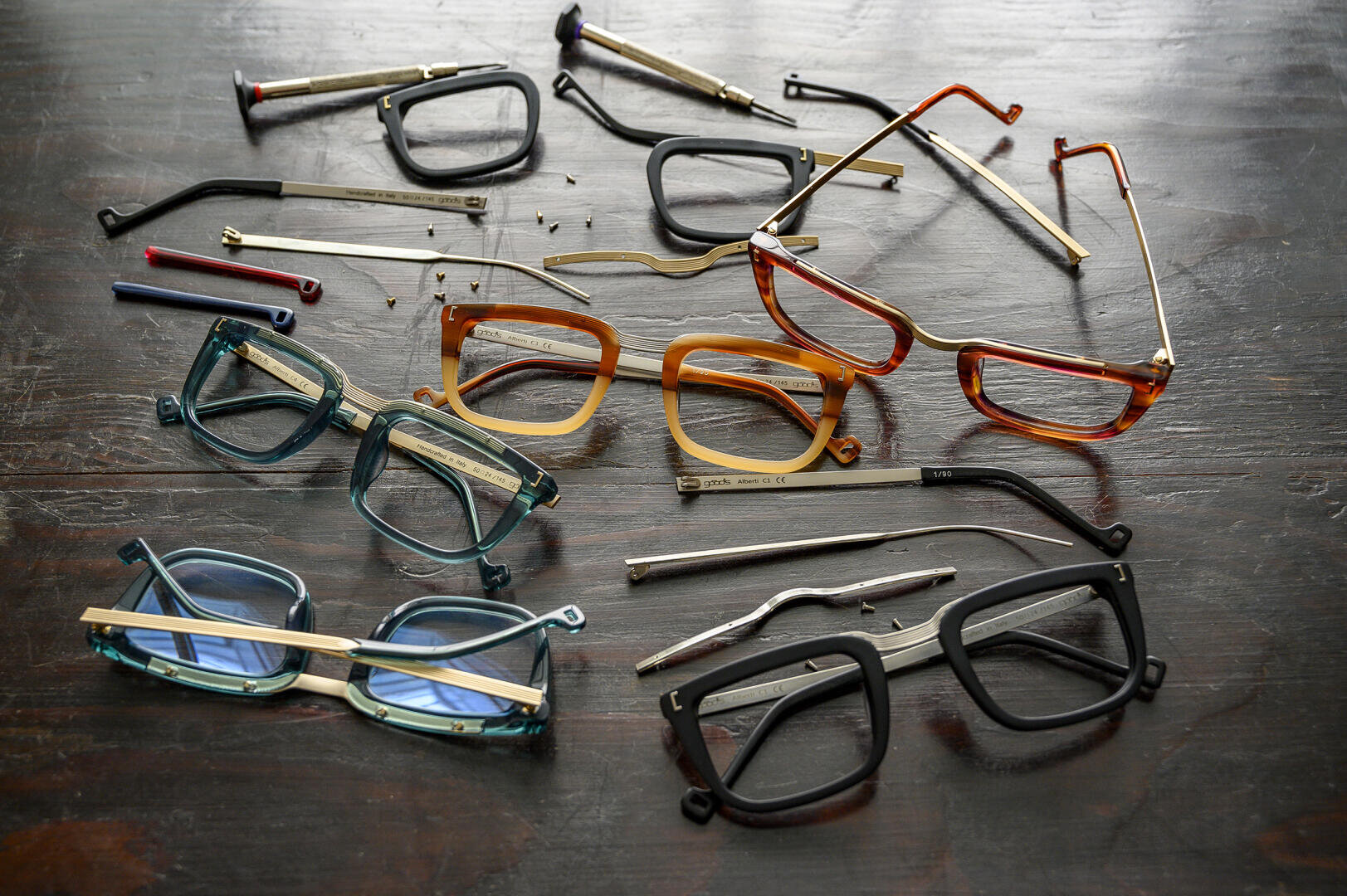 Mehrere Brillengestelle und -bügel in verschiedenen Farben und Stilen sind demontiert und auf einer dunklen Holzfläche ausgebreitet, wobei kleine Schrauben zwischen den Teilen verstreut sind.