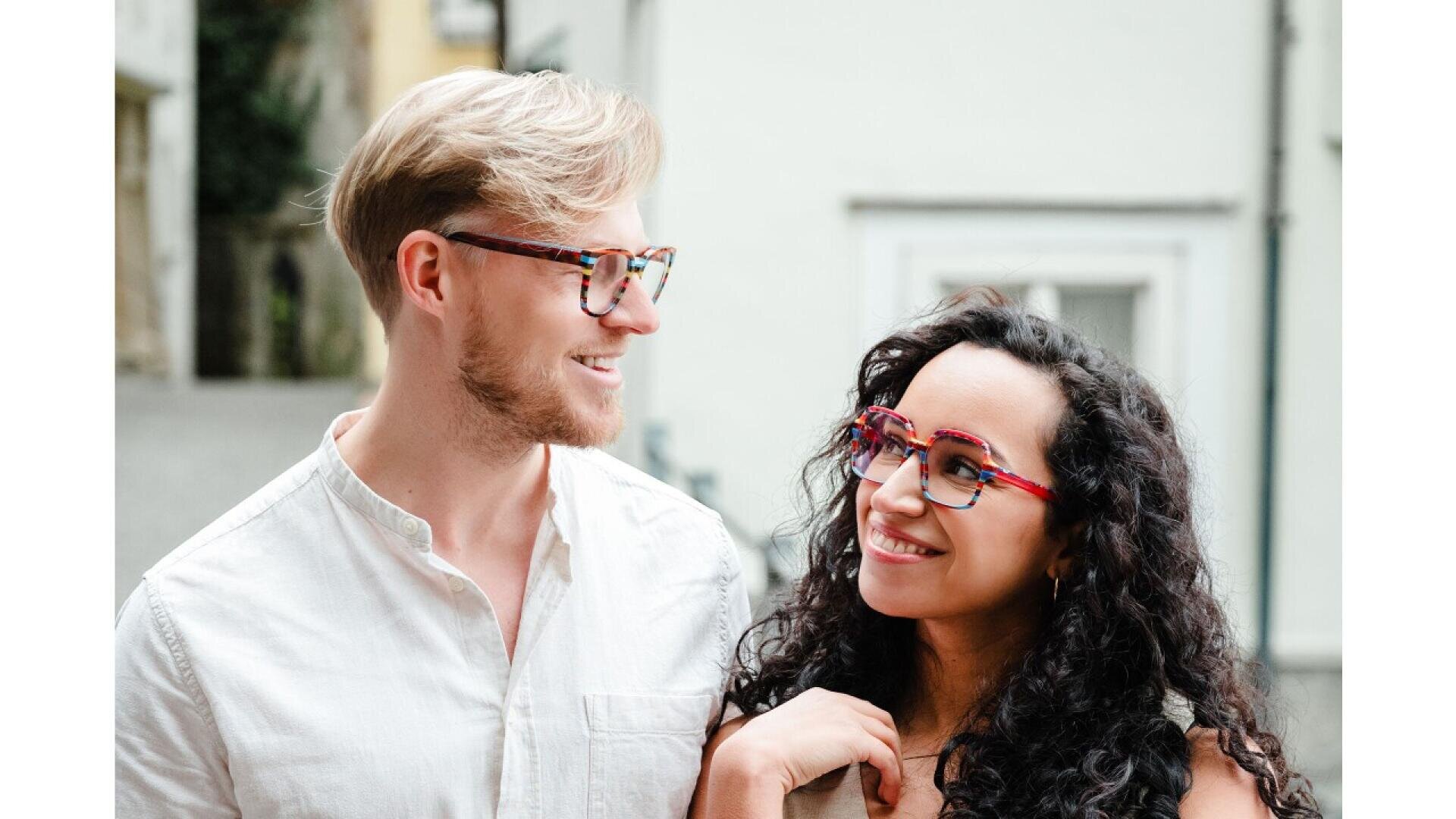 A man and woman wearing colorful eyeglasses smile at each other outdoors. The man has light hair and a beard, wearing a white shirt. The woman has long curly dark hair and is dressed casually. They appear happy and relaxed.