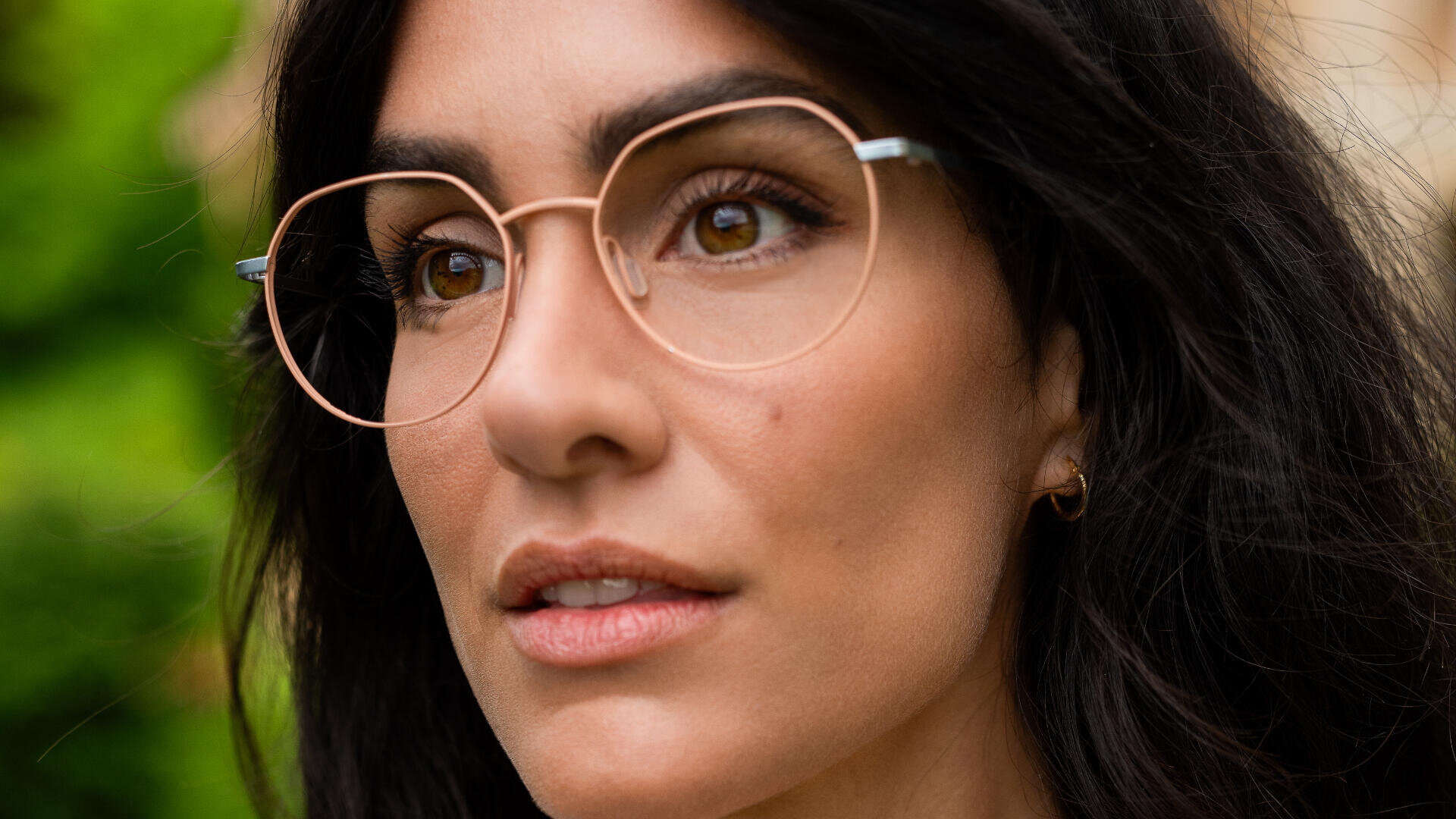 A close-up of a woman with long dark hair wearing thin, round, gold eyeglasses and a small gold hoop earring, looking slightly upwards. The background is blurred greenery.