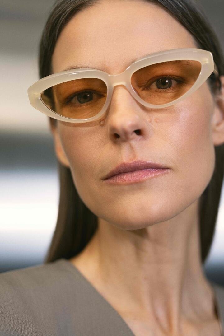 Eine Frau mit glattem braunem Haar trägt eine beigefarbene Cat-Eye-Sonnenbrille mit braunen Gläsern und einem neutralen Gesichtsausdruck, bekleidet mit einem grauen Oberteil, vor einem sanft verschwommenen Hintergrund.