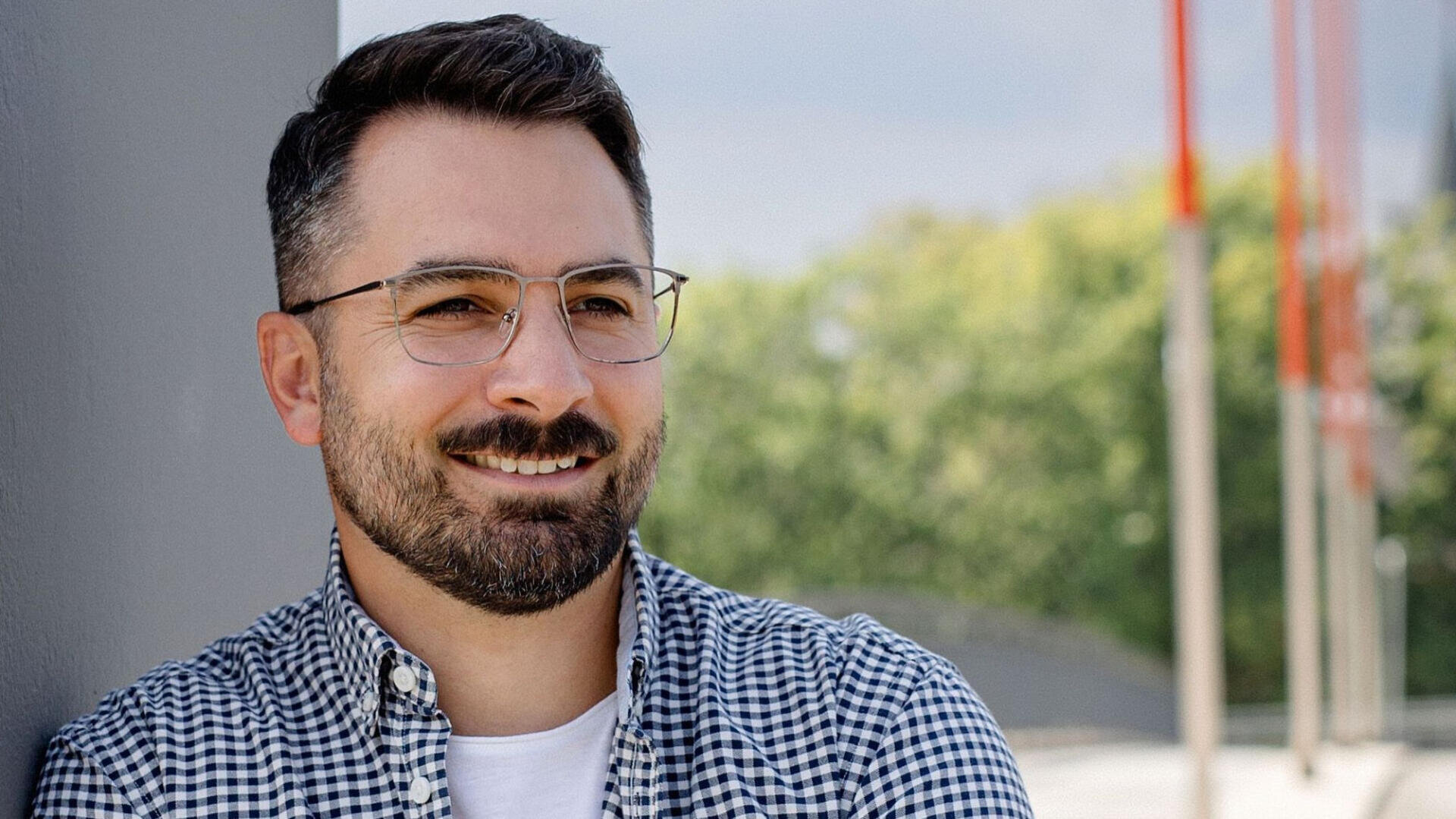 A man with short dark hair, a beard and glasses smiles as he leans against a wall outdoors. He is wearing a checked shirt over a white T-shirt, with trees and red pylons blurring in the background.