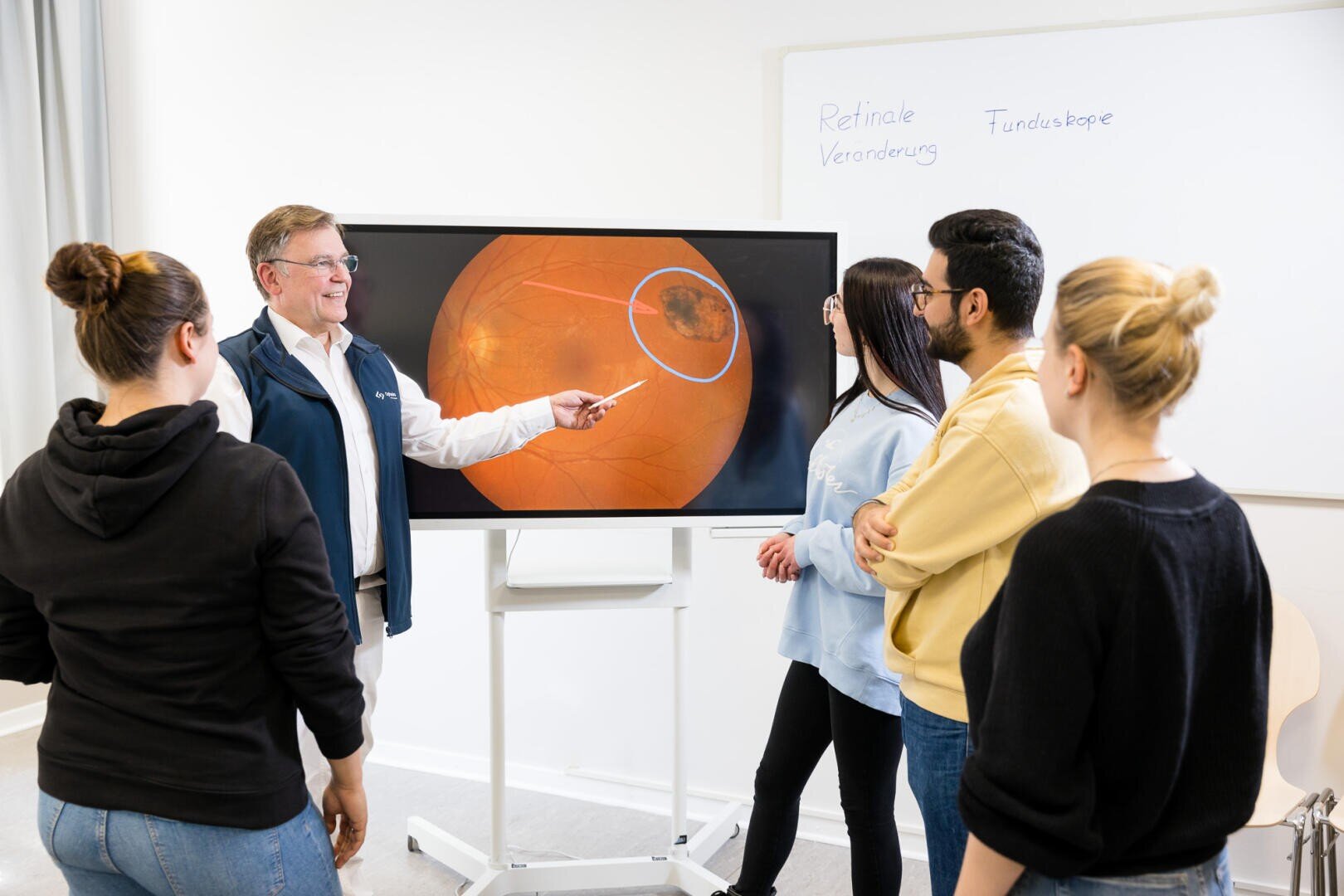 Ein Mann zeigt mit einem Zeiger auf einem großen Bildschirm auf ein medizinisches Bild eines Auges und erklärt es vier um ihn herum stehenden Personen. Auf einer Tafel im Hintergrund steht Retinale Veränderung und Funduskopie.