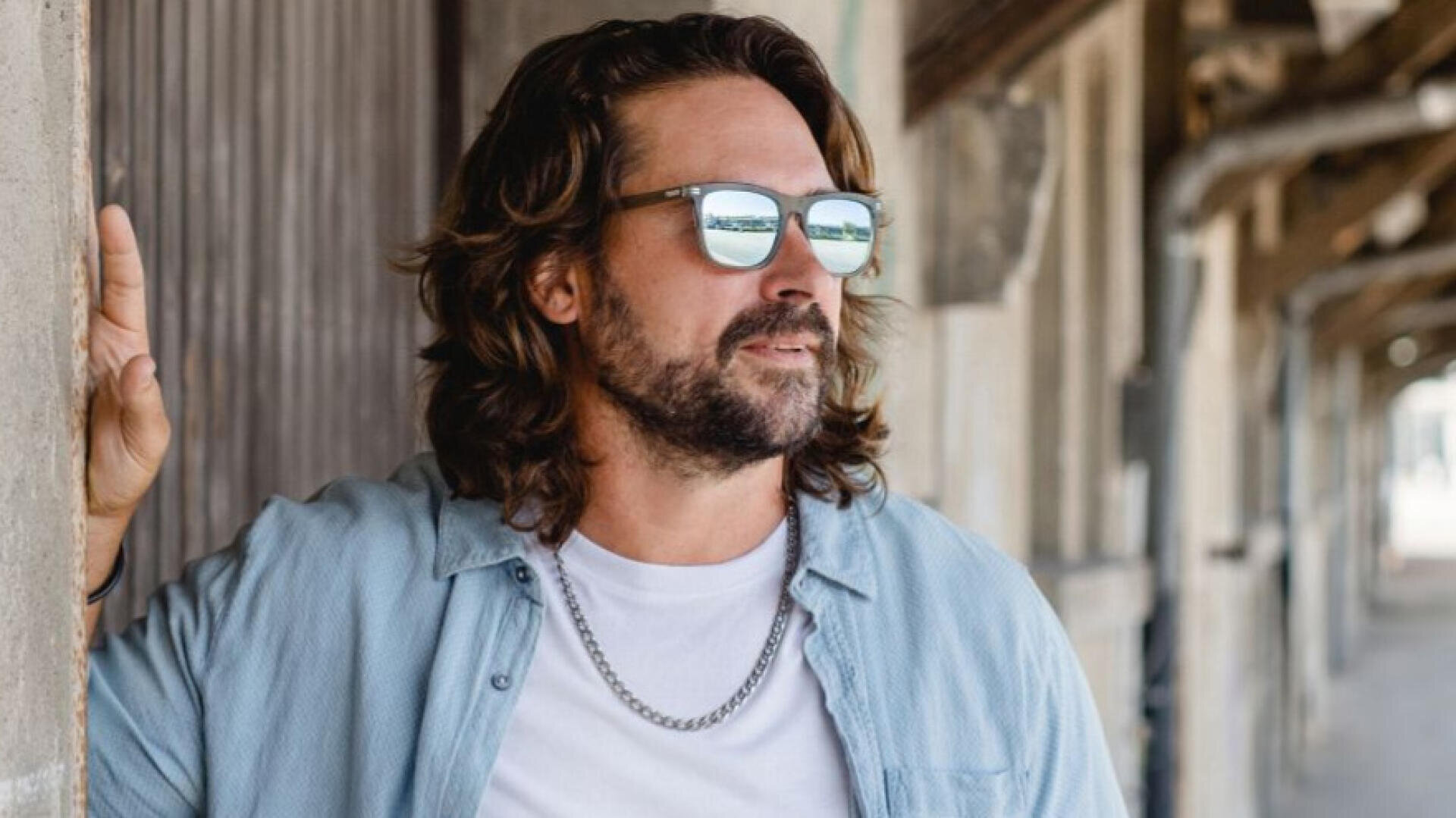 A man with long brown hair and a beard wears sunglasses, a light blue shirt over a white T-shirt and a silver necklace leaning against a concrete wall outside.