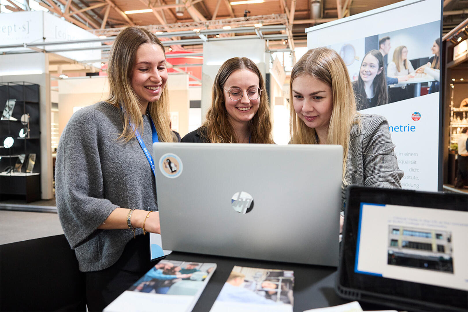Three young women are standing together, smiling and looking at a laptop on a table, which appears to be a professional event or conference. There are also flyers and a tablet on the table in front of them.