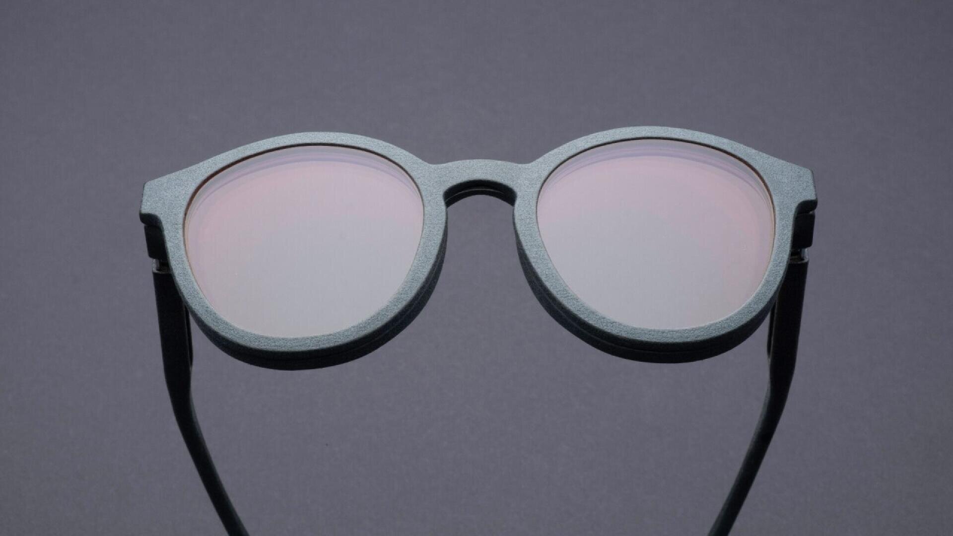 A pair of round, gray-framed sunglasses with pale pink-tinted lenses is displayed on a plain gray background.