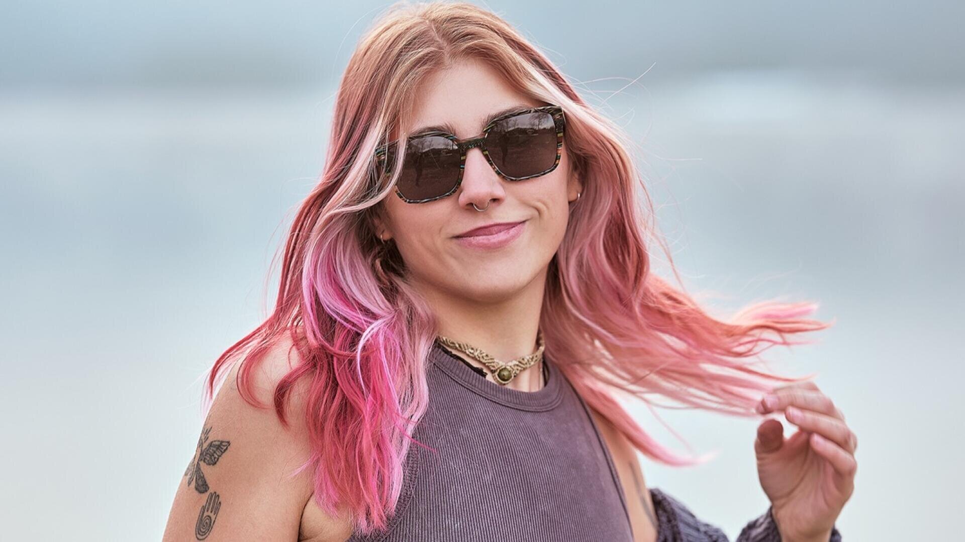 A young woman with long pink hair, wearing sunglasses and a sleeveless top, smiles at the camera. She has tattoos on her arm, a nose ring and is standing outdoors in front of a blurred background.