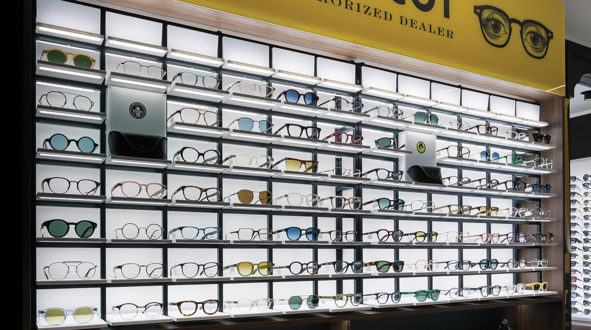 Eine Ausstellungswand in einem Brillengeschäft mit Dutzenden von Brillen und Sonnenbrillen in verschiedenen Formen, Farben und Stilen, die übersichtlich in mehreren Reihen beleuchteter Regale angeordnet sind.