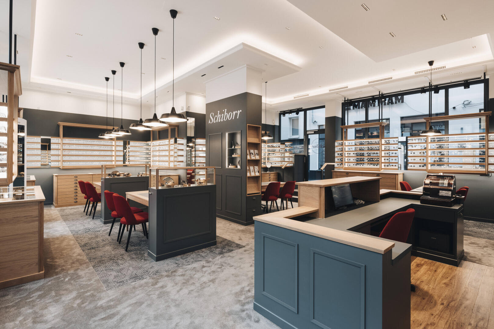 Modern eyewear store with wooden and blue-grey furniture, shelves filled with glasses, red chairs at tables, pendant lights and large windows that let in natural light.