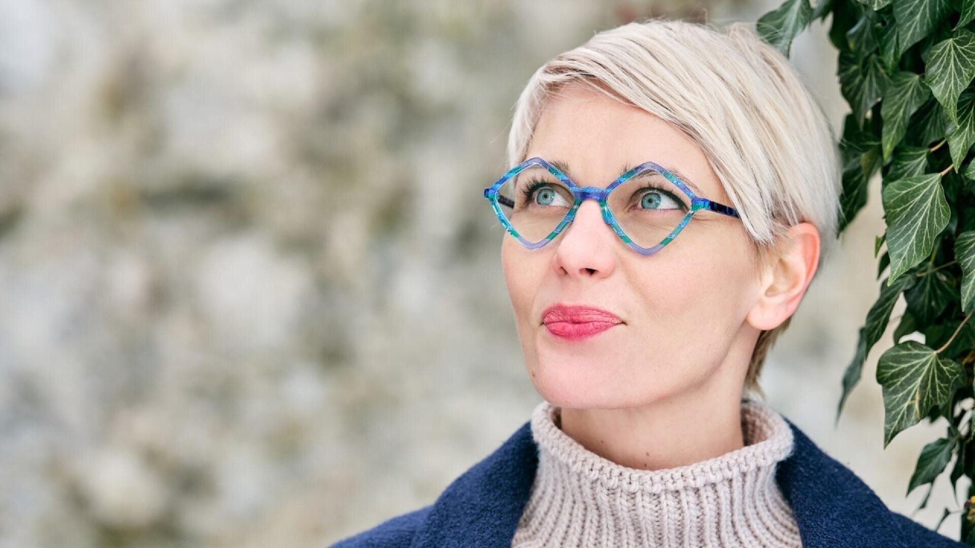 A woman with short blonde hair and blue geometric glasses looks upward. She is wearing a beige turtleneck sweater and a dark coat, standing next to green ivy against a blurred background.