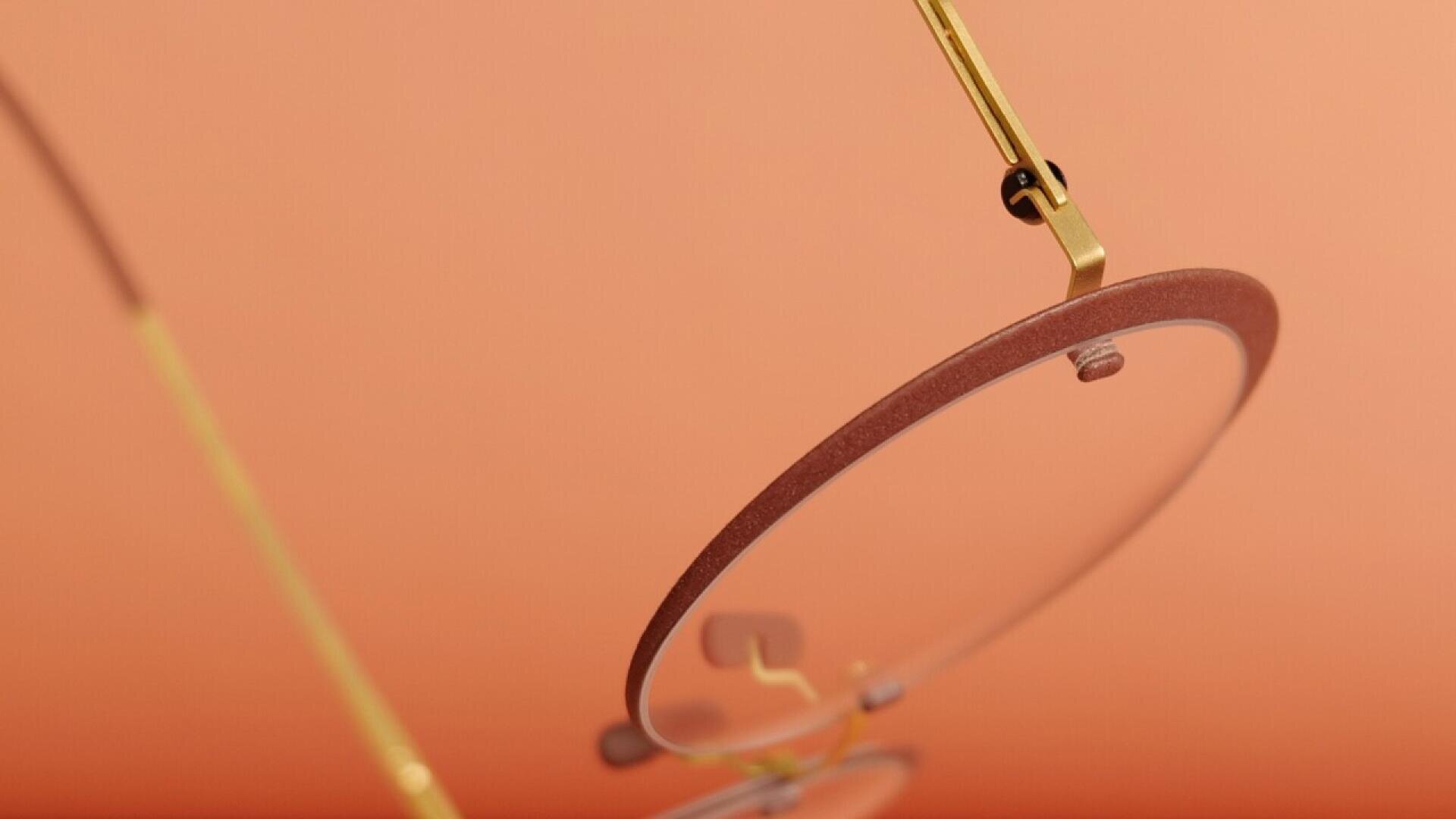 Close-up view of gold-framed eyeglasses with round lenses, set against a smooth peach-colored background. The focus is on the frames details, with one lens and part of the temple visible.