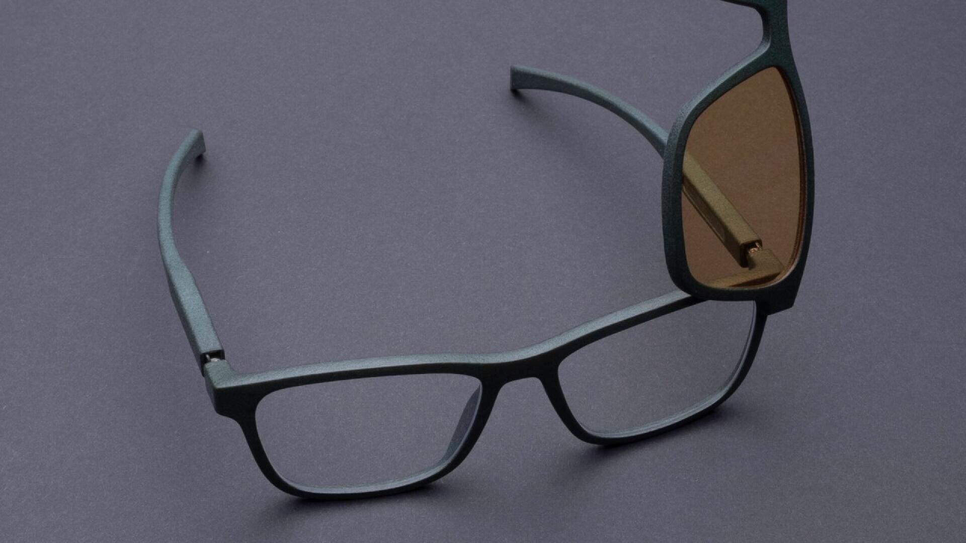 A pair of black rectangular eyeglasses with a brown-tinted clip-on lens partially attached, set on a dark gray surface.