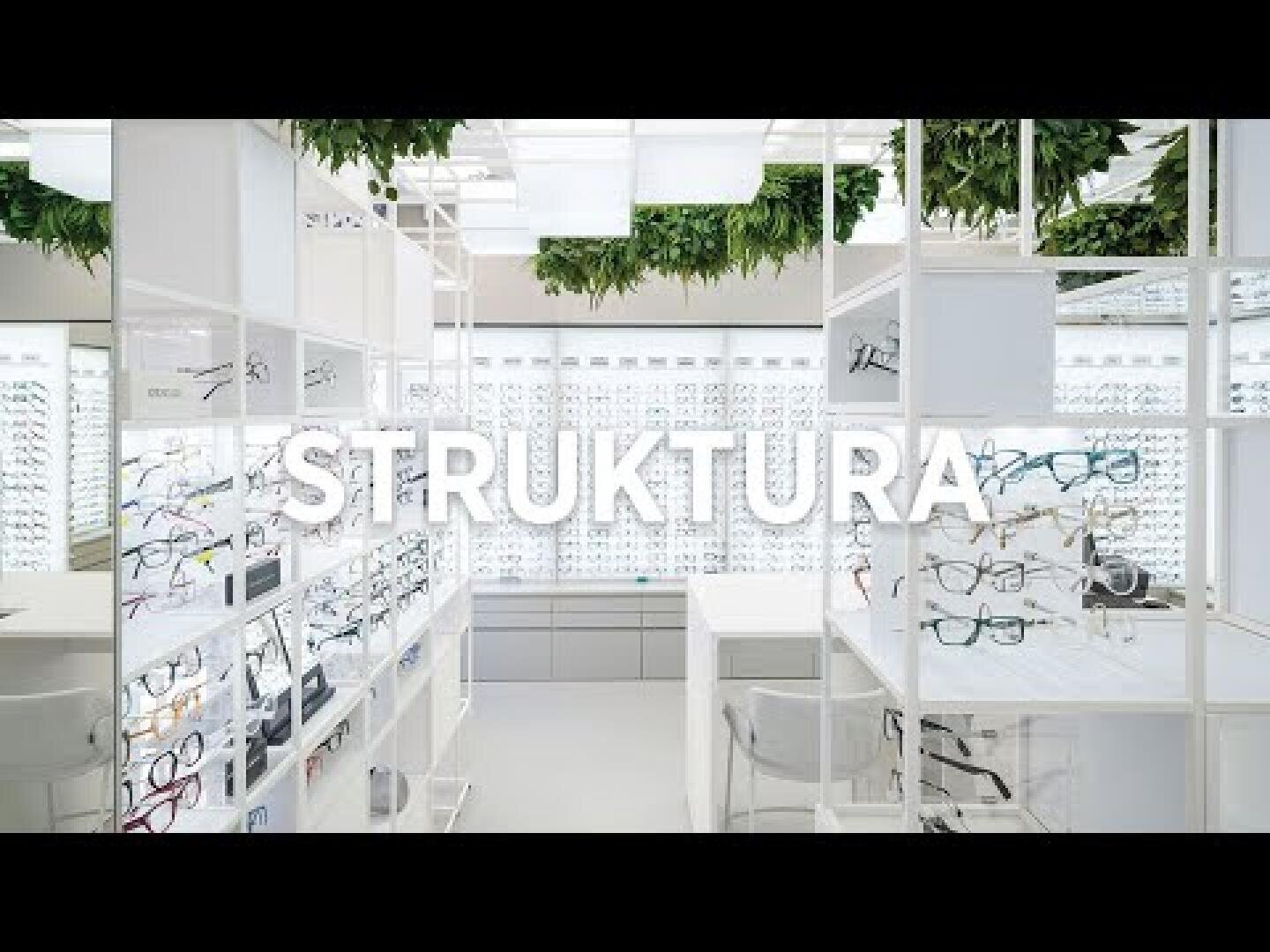 Modern eyewear store interior with white shelves displaying various eyeglasses. The word STRUKTURA is overlaid in large text. Green plants hang from the ceiling, adding a natural touch to the clean, bright space.