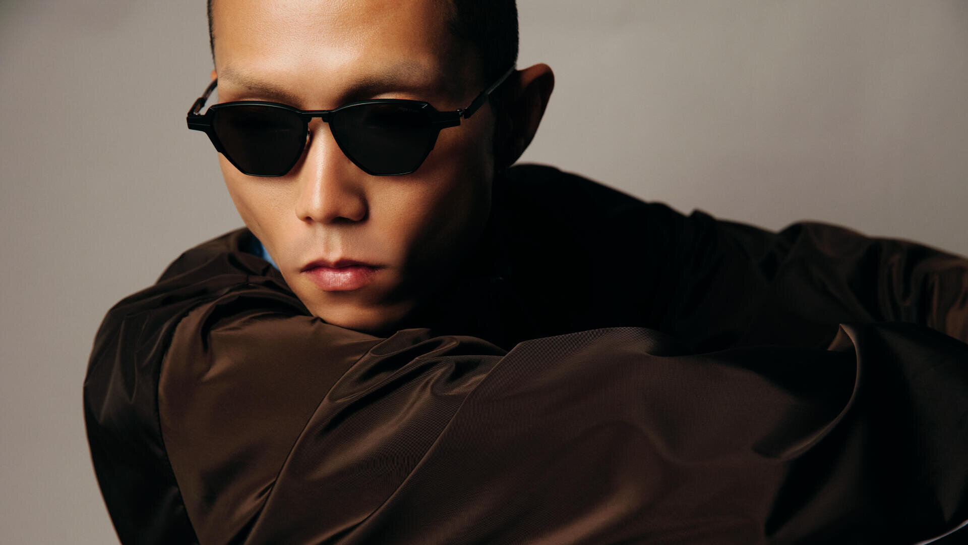 A person wearing black sunglasses and a dark, shiny jacket looks away from the camera with a serious expression against a neutral background.