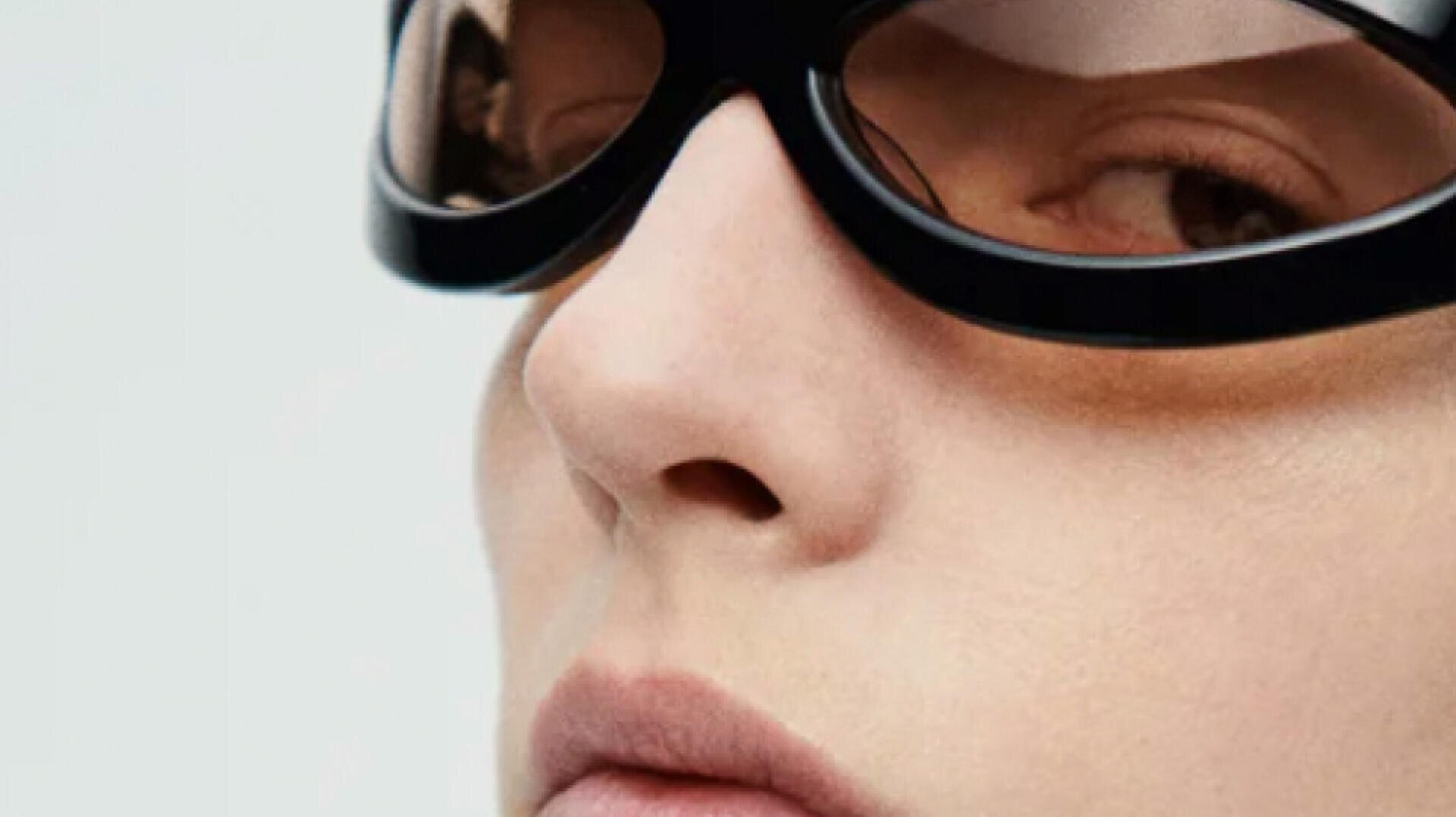 Close-up of a persons face wearing large, black sunglasses with brown lenses. The focus is on the nose, lips, and one eye, with soft lighting and a neutral background.